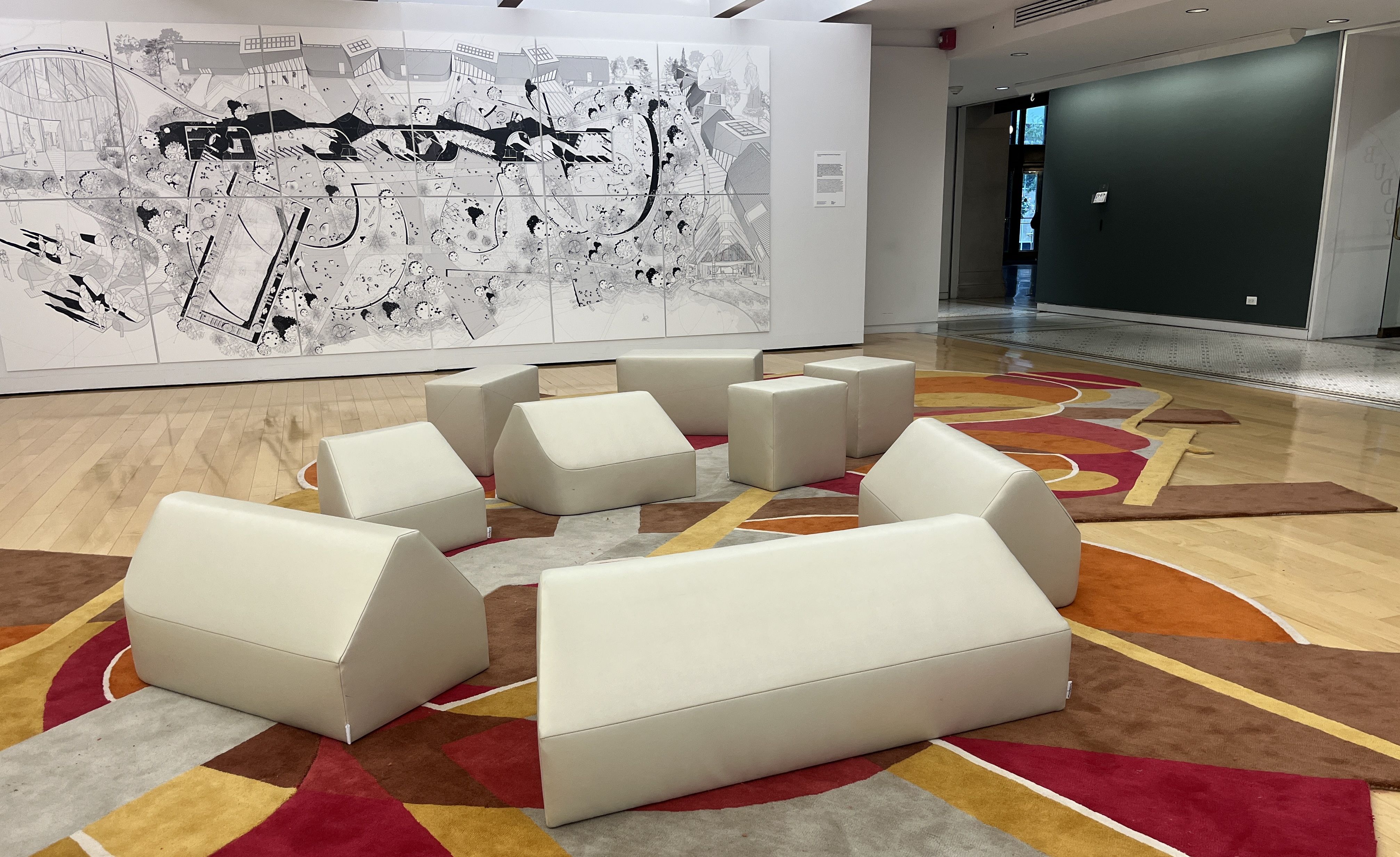 Chicago Cultural Center gallery with a large black-and-white abstract mural on the wall and a colorful geometric rug on the wooden floor. Beige geometric soft seating pieces arranged on the rug.