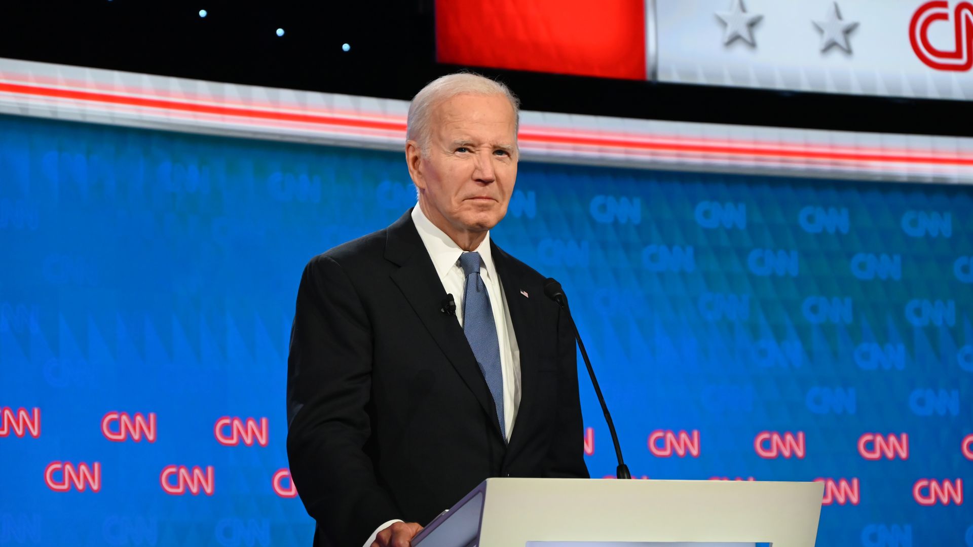 President Biden in a dark suit standing behind a white podium.
