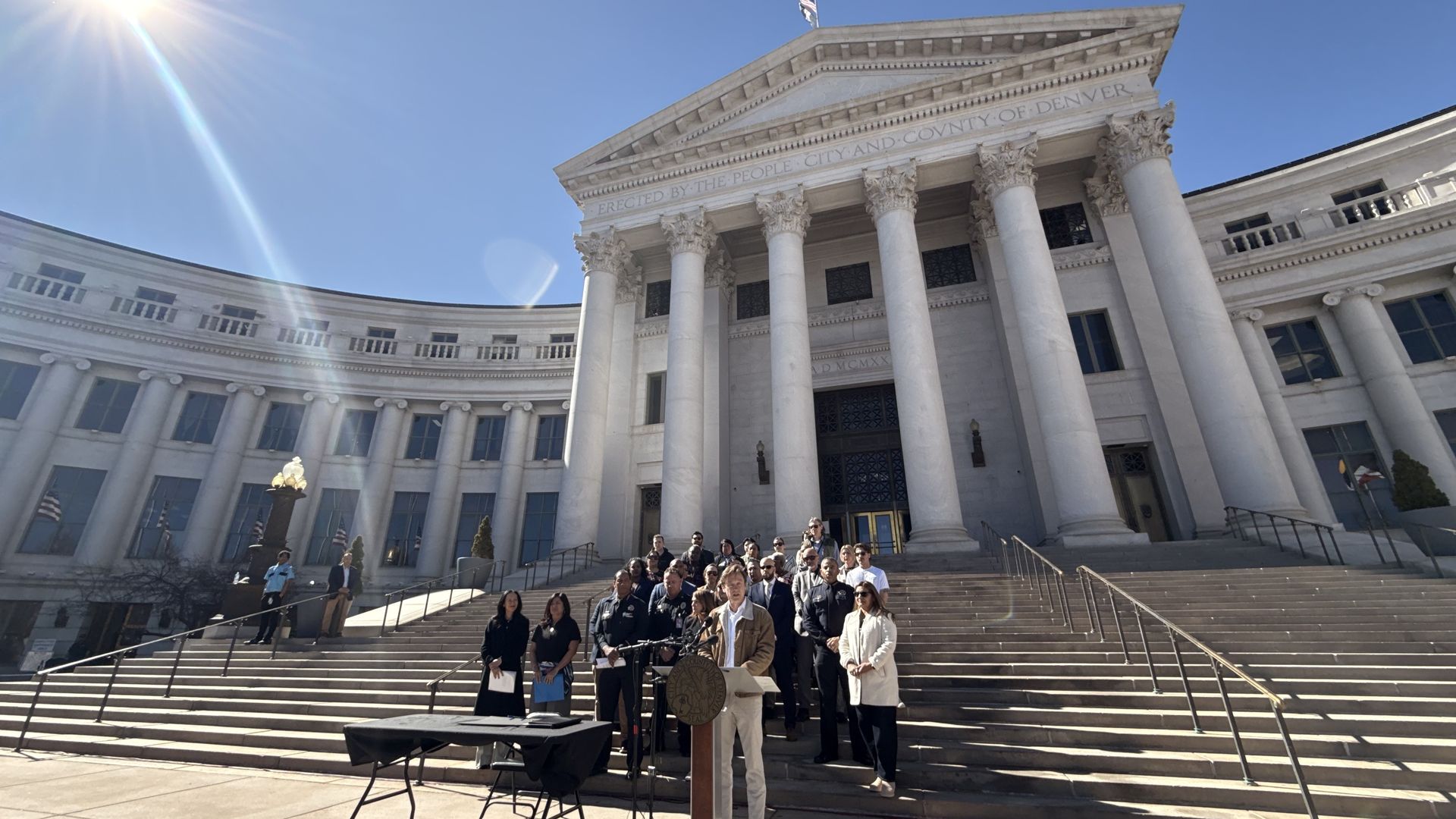Group of people, including a speaker at a podium, gathered on steps of a large white government building under bright sun with blue sky and lens flare.