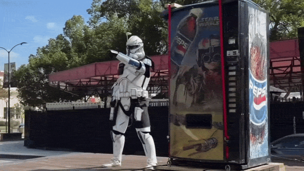 A stormtropper next to a Star Wars vending machine moving on a flat bed truck with red bleachers in the background. 