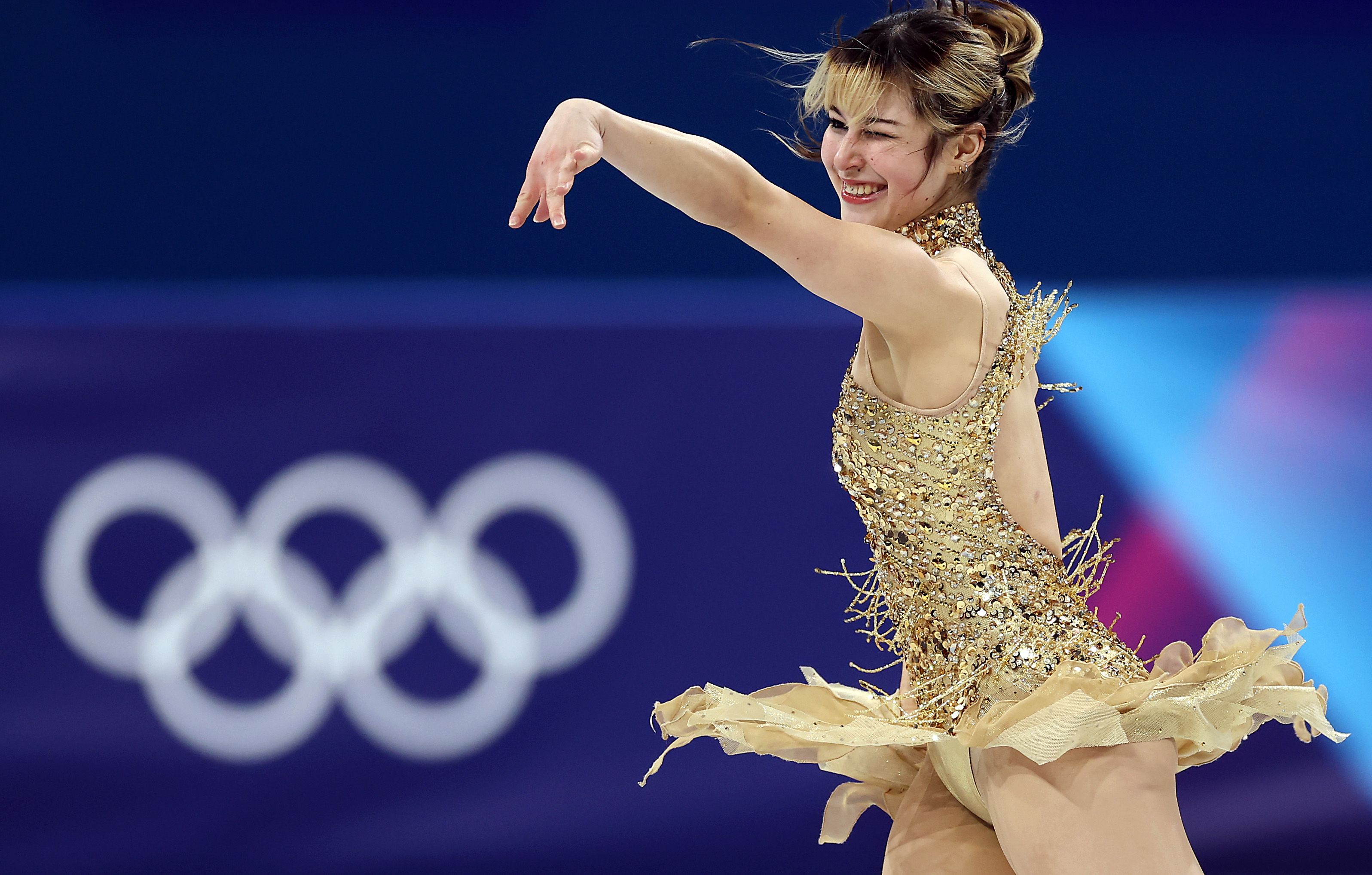 Figure skater in a sparkling gold costume performs a spin with a winking smile on her face against a background featuring blurred Olympic rings.