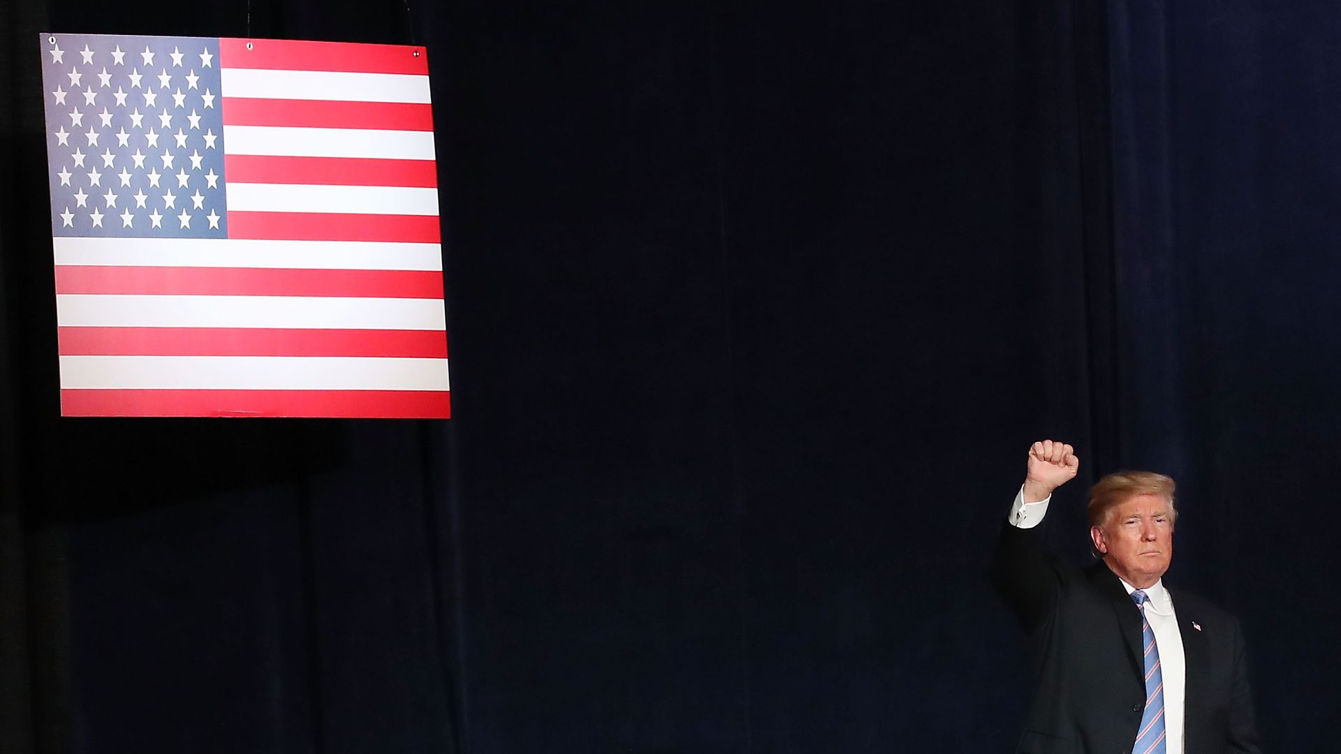 Donald Trump raises his arm in a fist before a navy blue background and an American flag.