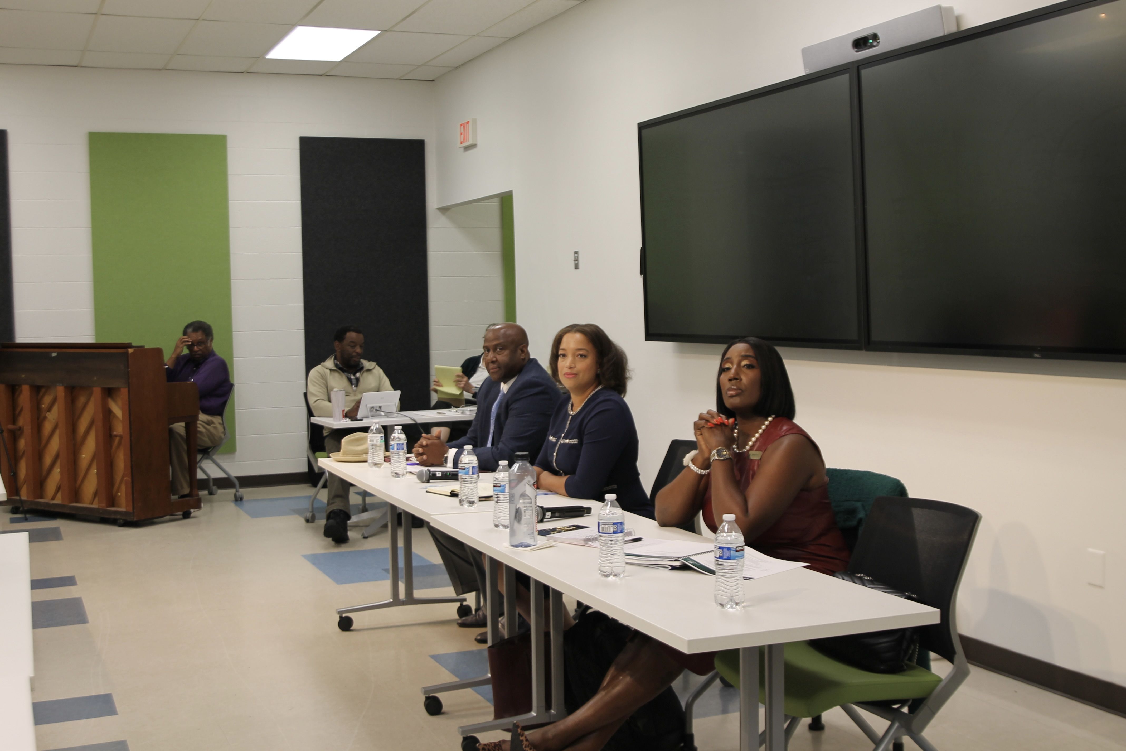 Panel of three people seated at a table with water bottles and papers in a conference room with two large blank screens on the wall and a piano in the background.