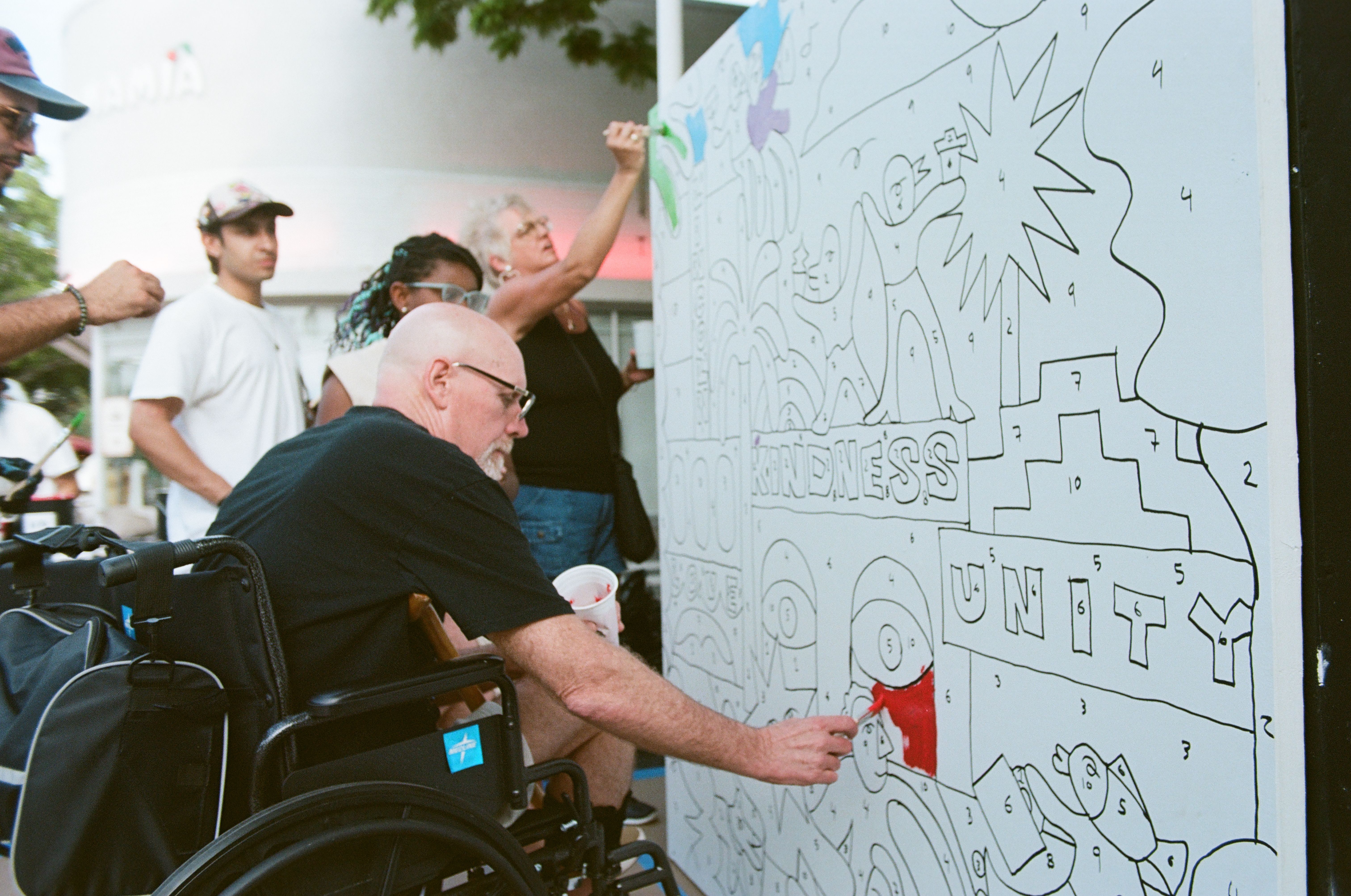 Outdoor scene with a tall white wall covered in a large black-and-white doodle maze. A bald man in a wheelchair holds a marker as others look on and one person adds color.