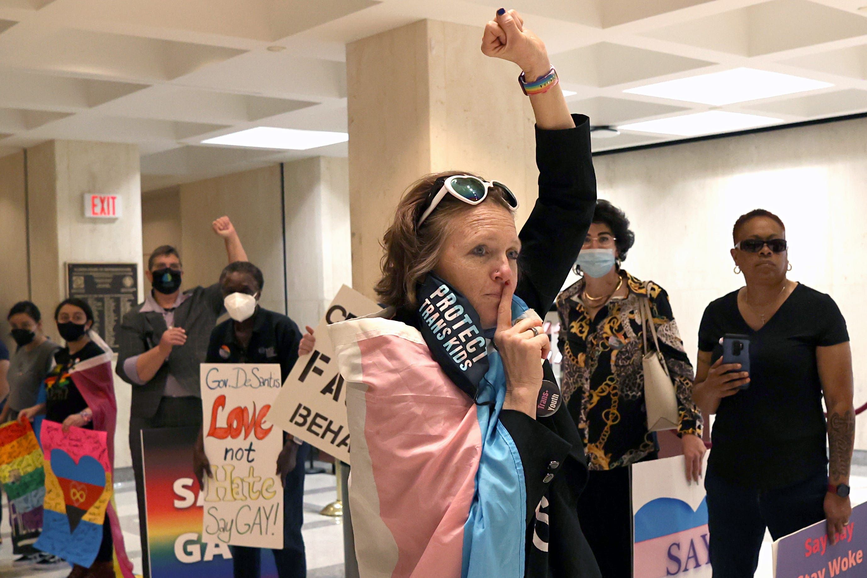 A person wrapped in the trans flag holds a finger to their lips and their fist in the air.