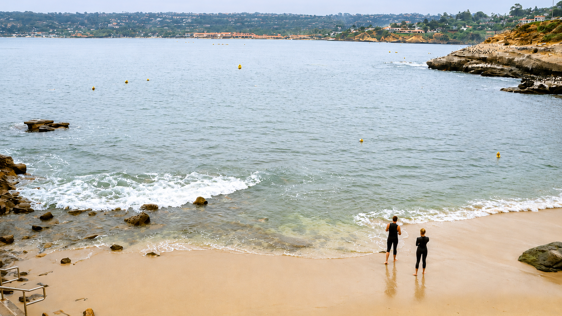 Two people in wetsuits stand on a sandy beach near the water while gentle waves roll in. Rocky outcrops frame the shore; a distant town on green hills lines the horizon under a cloudy sky.