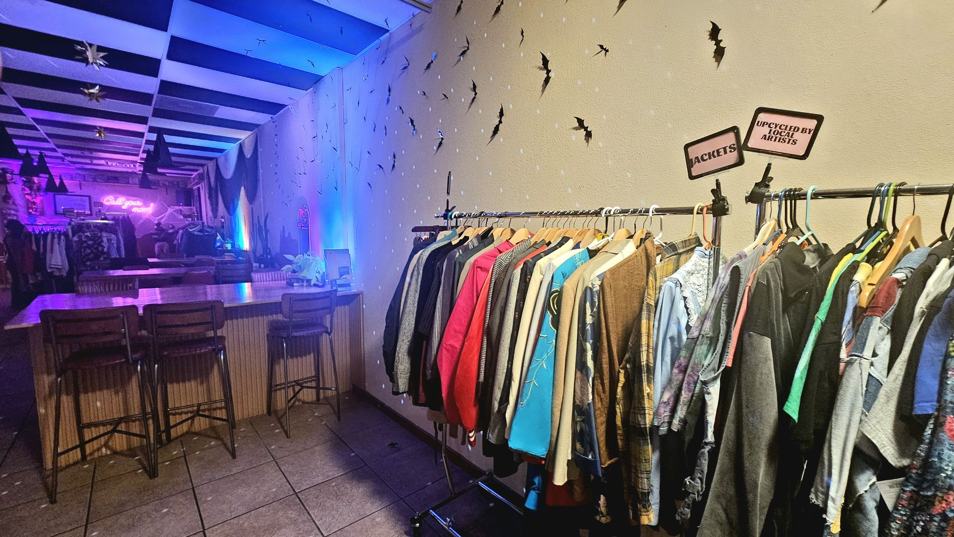 Dim, neon-lit shop with a long rack of colorful jackets, bat decals on the wall, and a neon "Call your mom" sign; a bar area with stools under blue-purple lighting.