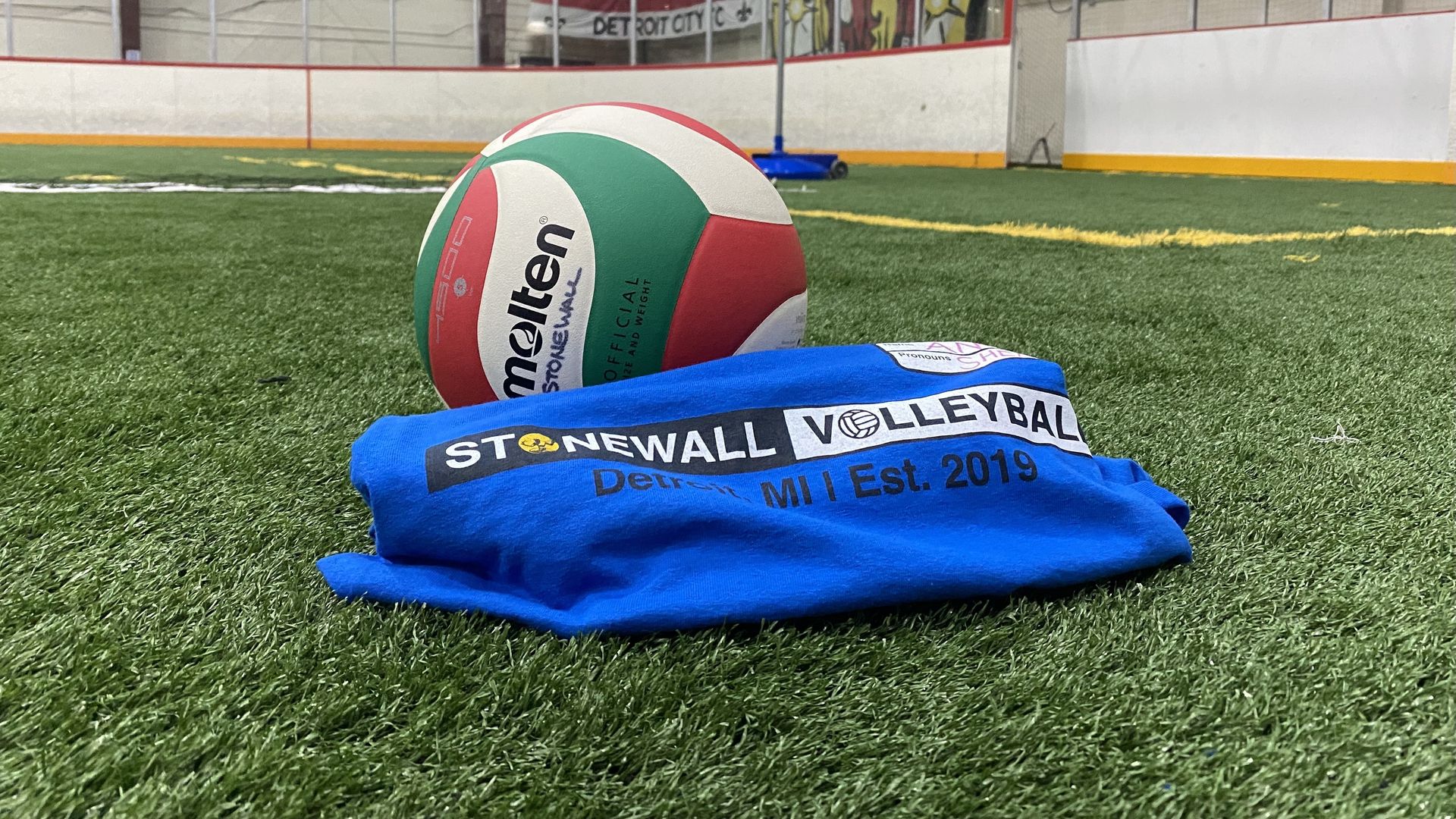 A volleyball sits next to a team t-shirt that says "Stonewall Volleyball" on a turf field.
