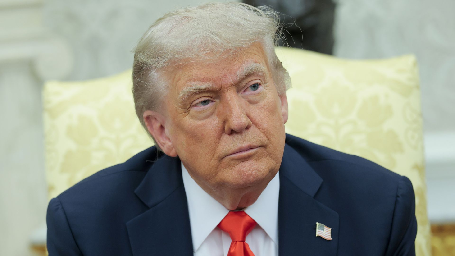 Close-up of Trump in the Oval Office with light blond hair wearing a dark blue suit, white shirt, and red tie, seated against a pale yellow floral patterned background.