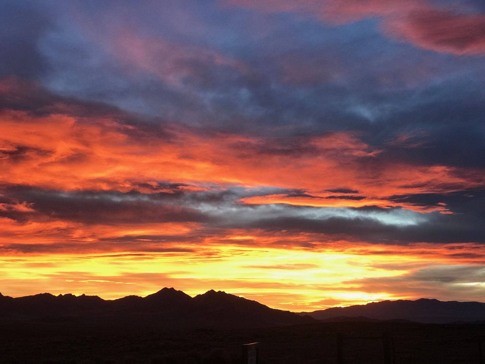 Sunset scene with fiery orange and red clouds across the lower sky, fading to purple and blue higher up; dark mountain silhouettes outline the horizon, with a dim foreground.