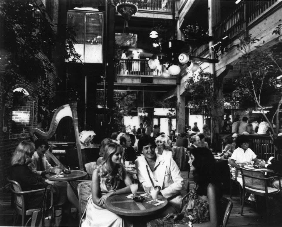 Black-and-white indoor courtyard cafe with round tables; foreground shows three friends—blonde woman, man in a light suit, and dark-haired woman—smiling as they chat. Background features plants and a two-level atrium with hanging lanterns.