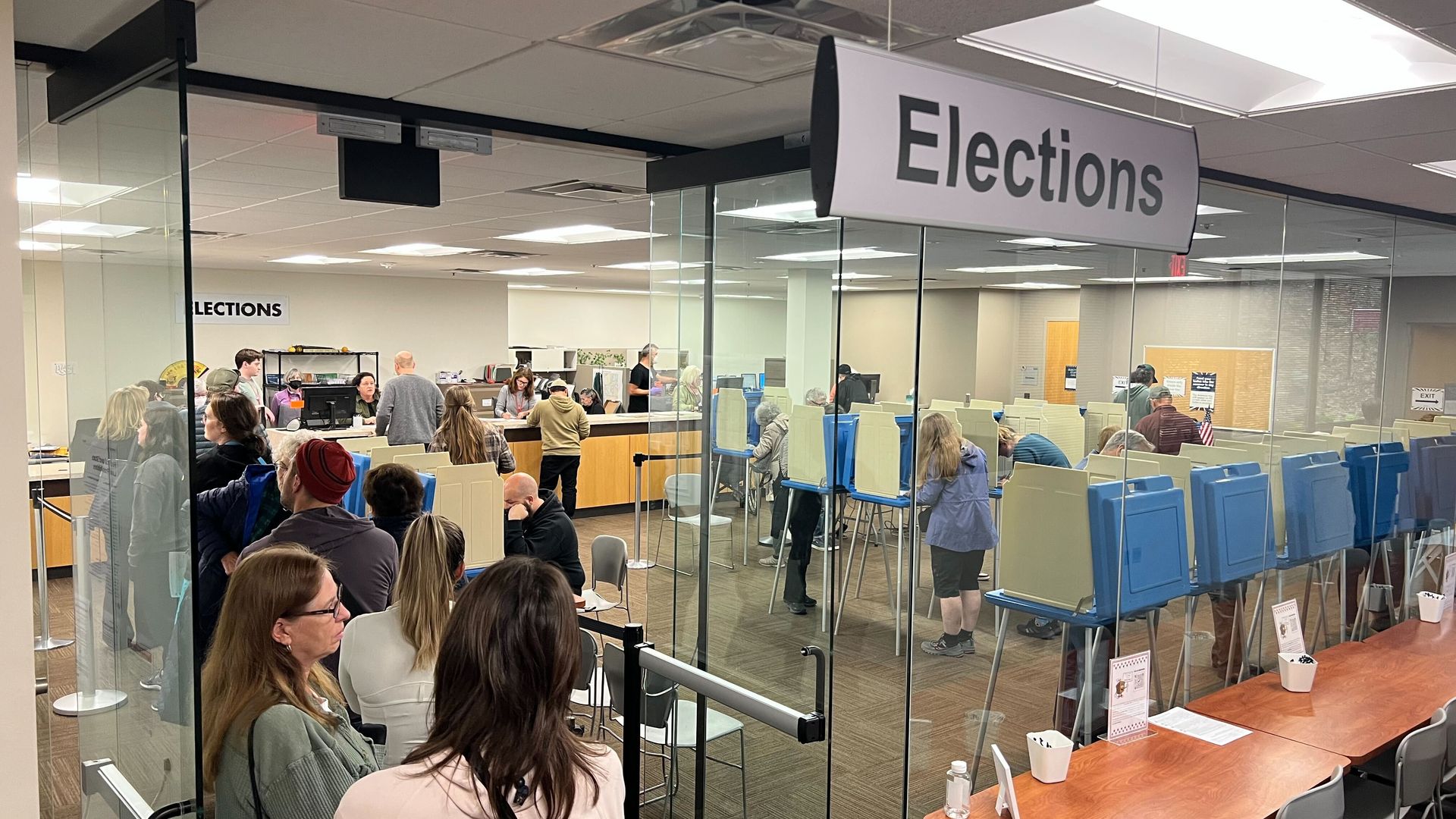 Voting booths in a glass-walled government office with a long line of people under a sign reading "Elections"