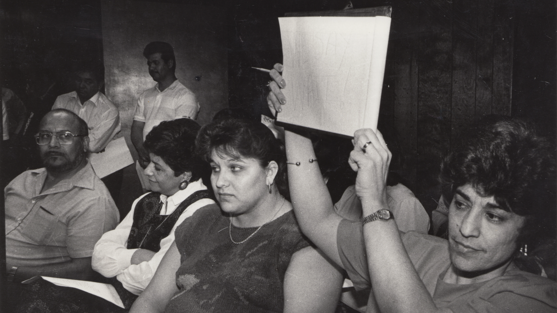 A black and white photo shows adults sitting at a meeting and woman holding up a sign, though the sign is not legible