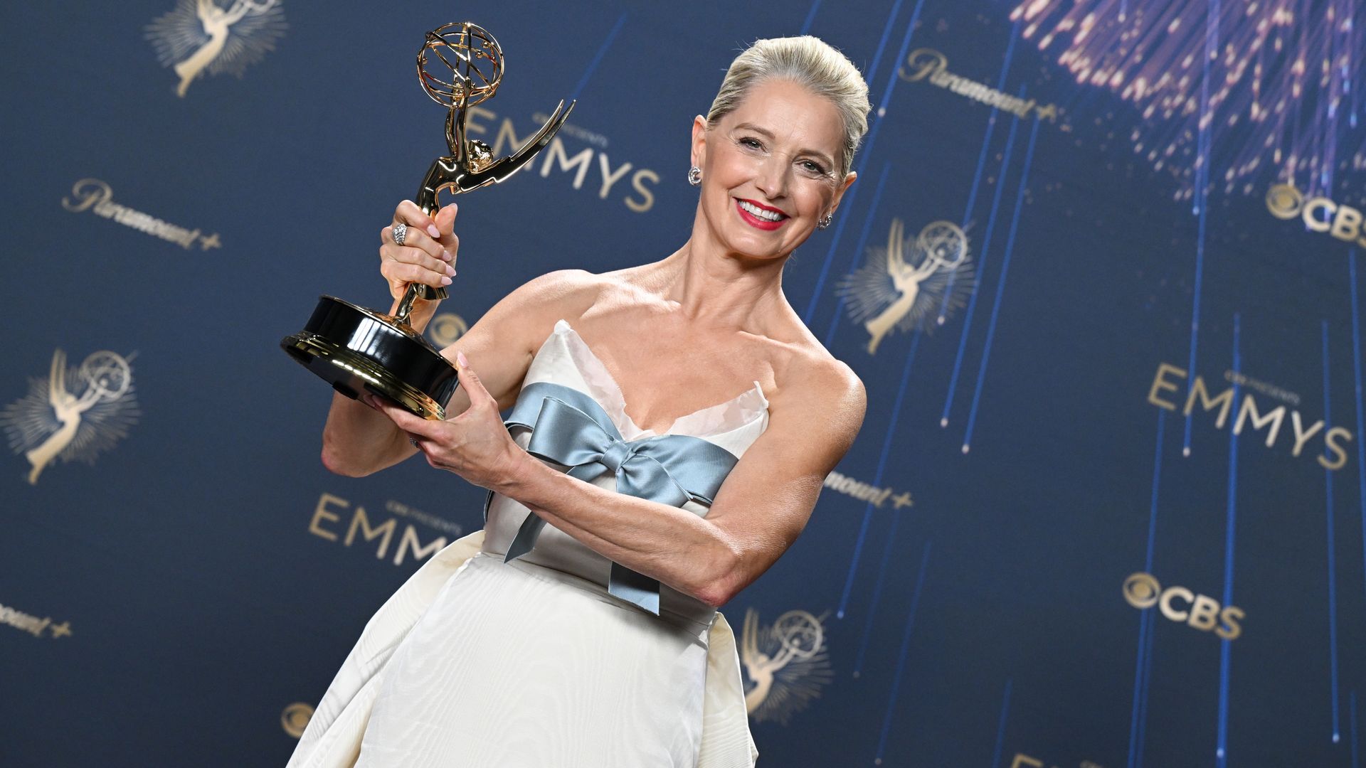 A woman wears a white gown with a blue bow across the chest and holds up an Emmy while she smiles at the camera. A wall behind her has CBS, Emmys and Paramount logos on it.