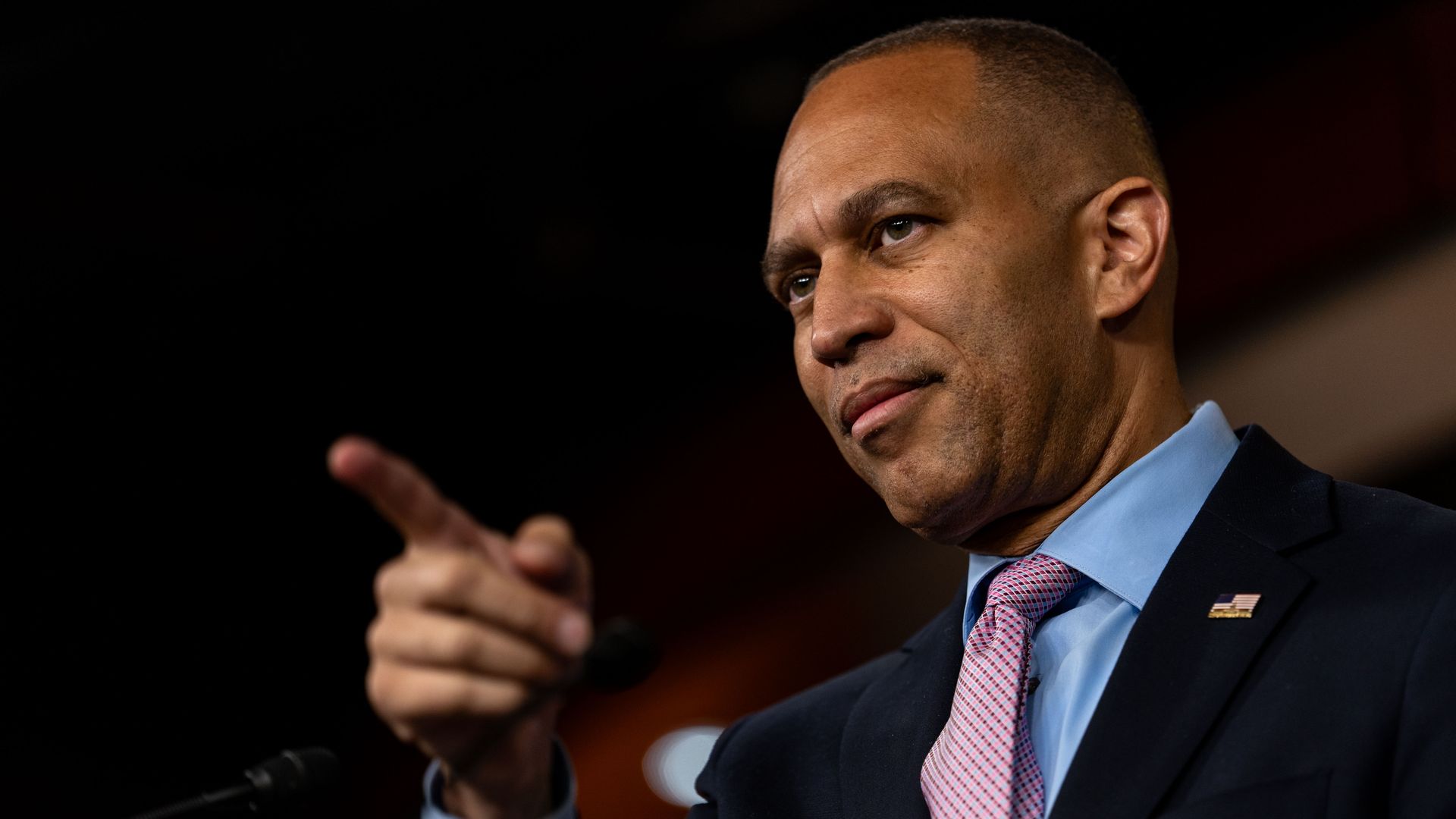 Hakeem Jeffries, wearing a gray suit, points while standing in a dimly lit, wood-paneled room.