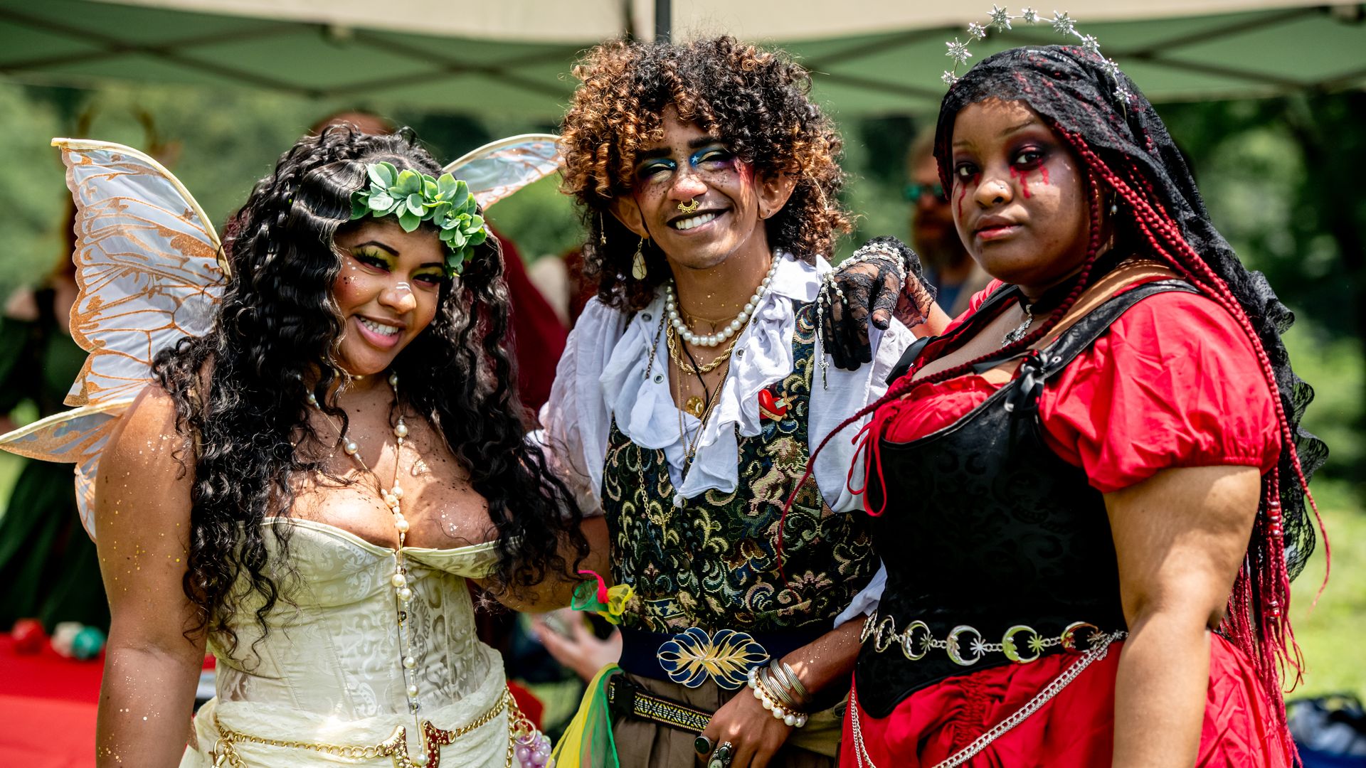 Three costumed people pose outdoors under a canopy. Left: cream gown with butterfly wings and a green leaf crown. Center: curly hair and blue makeup. Right: red and black outfit with a star headpiece.