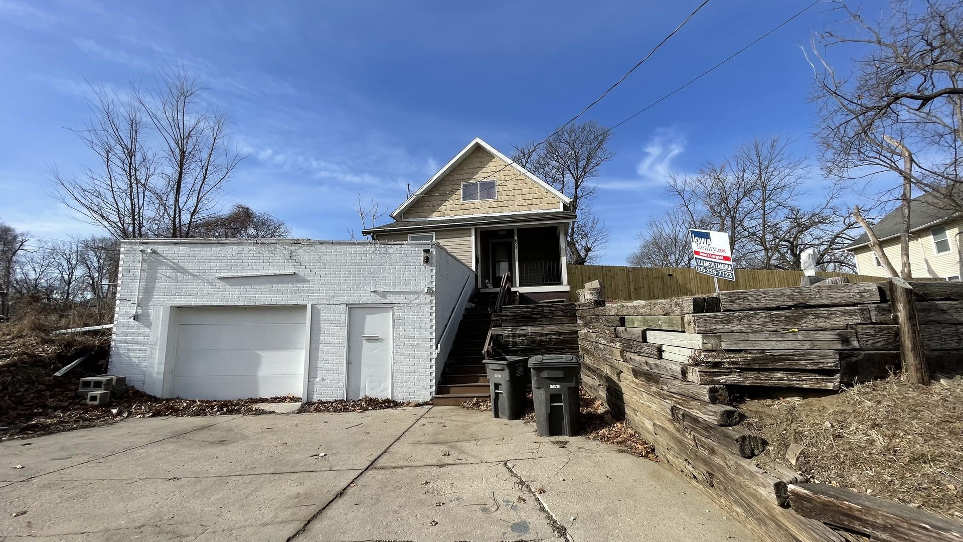 Small beige house with front porch on hill, white brick garage below with driveway, wooden retaining wall on right, leafless trees, clear blue sky, realty sign visible.