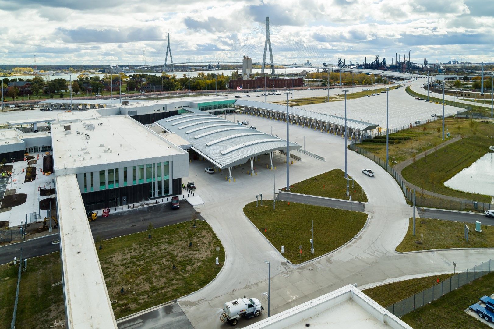 A large modern border checkpoint facility with multiple covered lanes, adjacent parking, surrounded by grassy areas and a distant cable-stayed bridge.