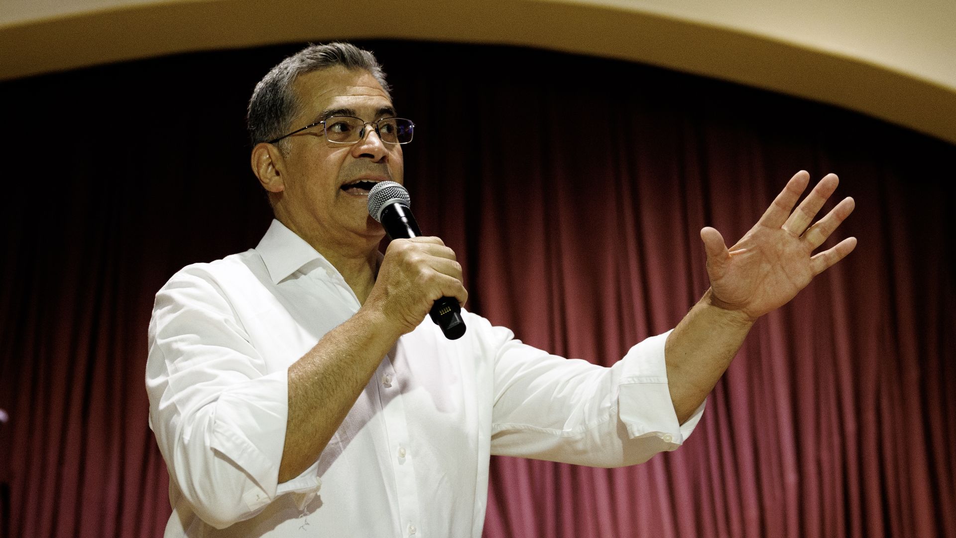A middle-aged man wearing a white shirt and glasses speaks into a handheld microphone on stage, his left hand raised in a gesturing motion, with maroon curtains in the background.