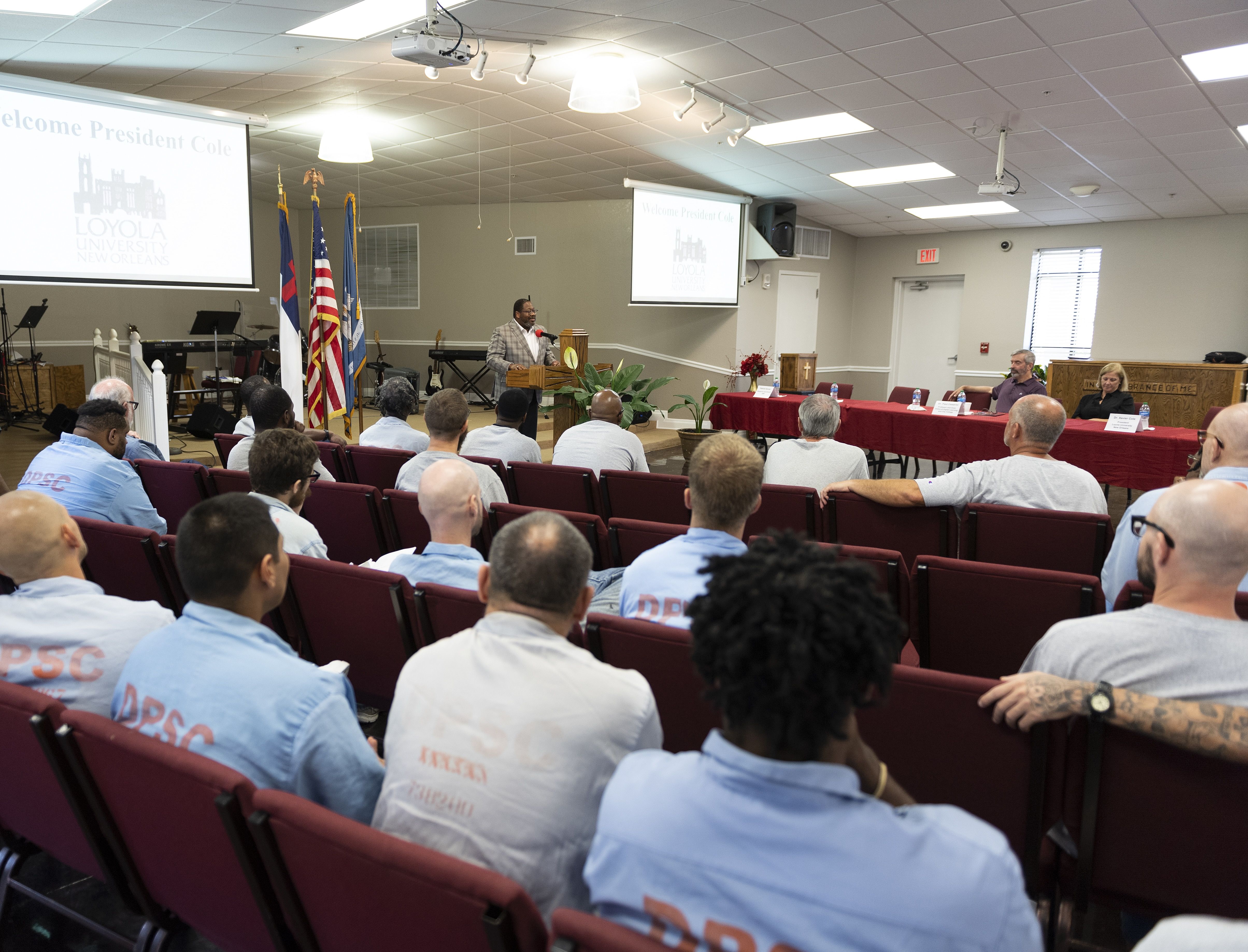 Man in plaid jacket speaking at podium in front of an audience wearing light blue DPSC shirts, seated in maroon chairs in a room with Loyola University New Orleans slides and flags displayed.