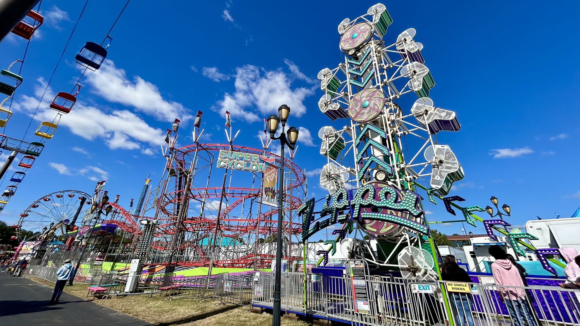 Bright blue sky above a lively amusement park with colorful chairlift, red roller coaster labeled "Super Cyclone," and a spinning ride with green and purple seats and neon lights.