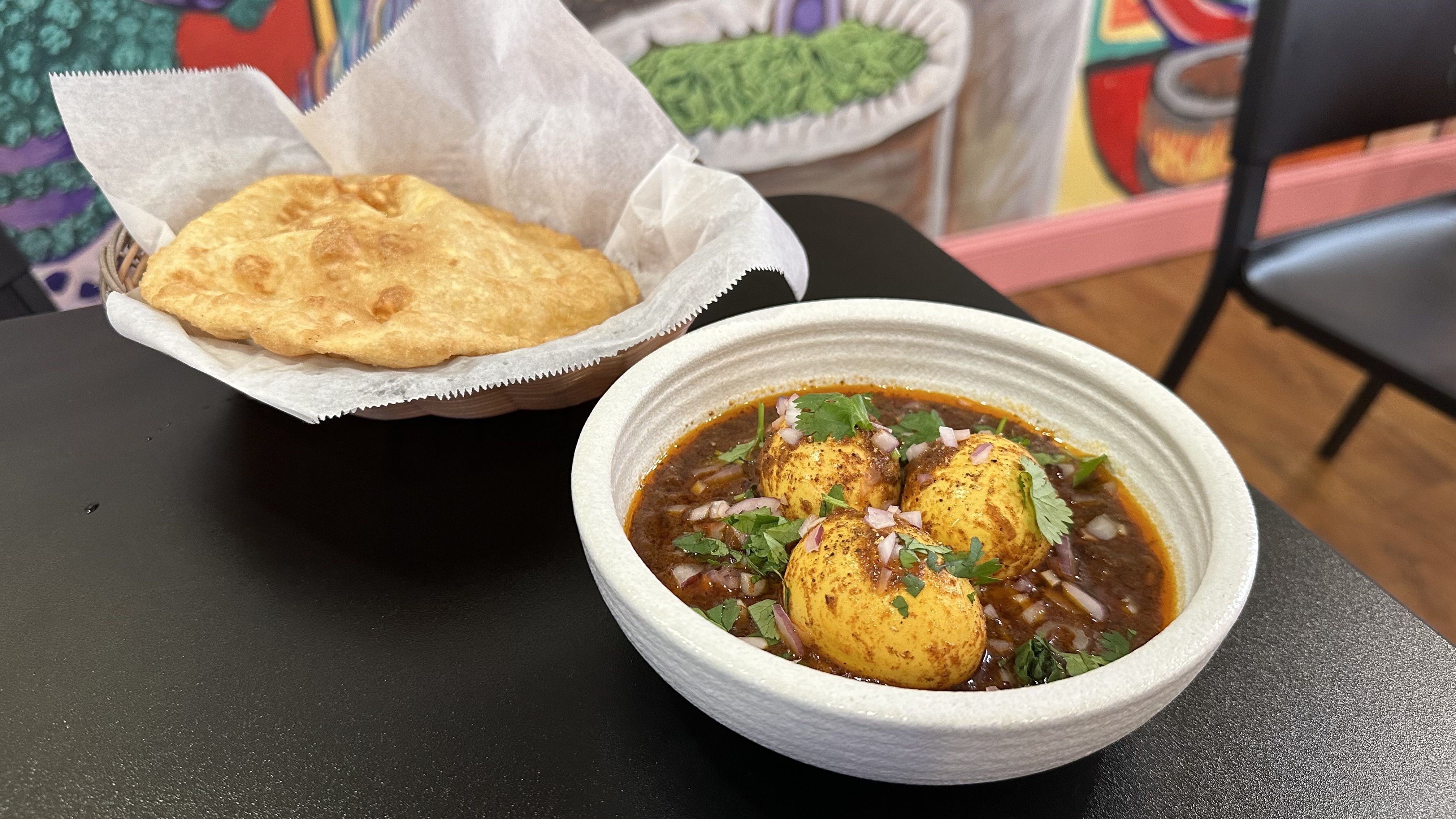 A bowl of curry filled with three hard-boiled eggs sits on a table next to a basket with a piece of fried bread in it.