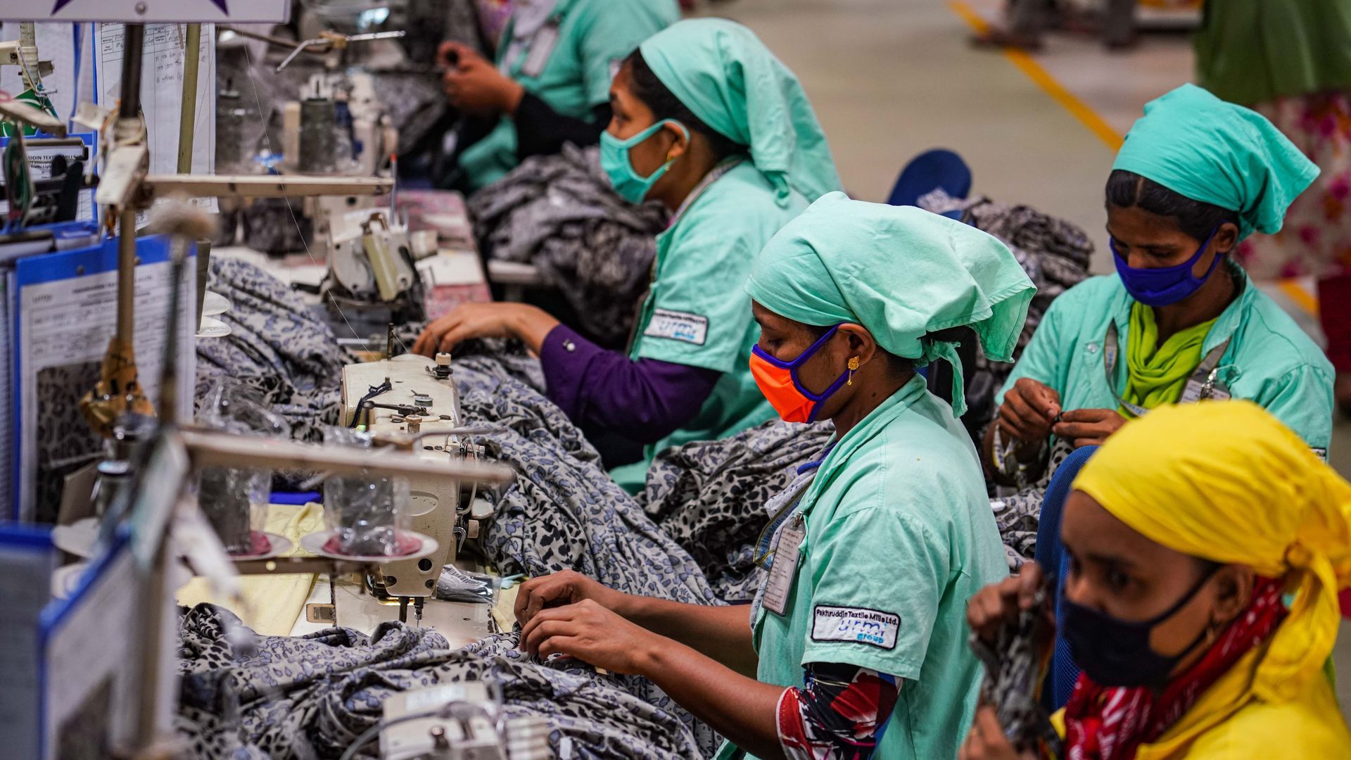Photo of workers wearing light aqua outfits and masks sewing clothing together in a factory
