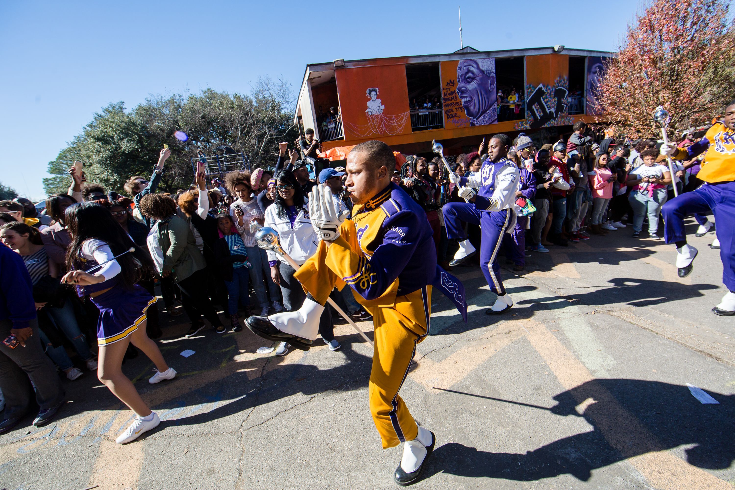 Marching band members in purple and gold uniforms perform high-stepping moves on a street, surrounded by a crowd of spectators taking photos and watching on a sunny day.