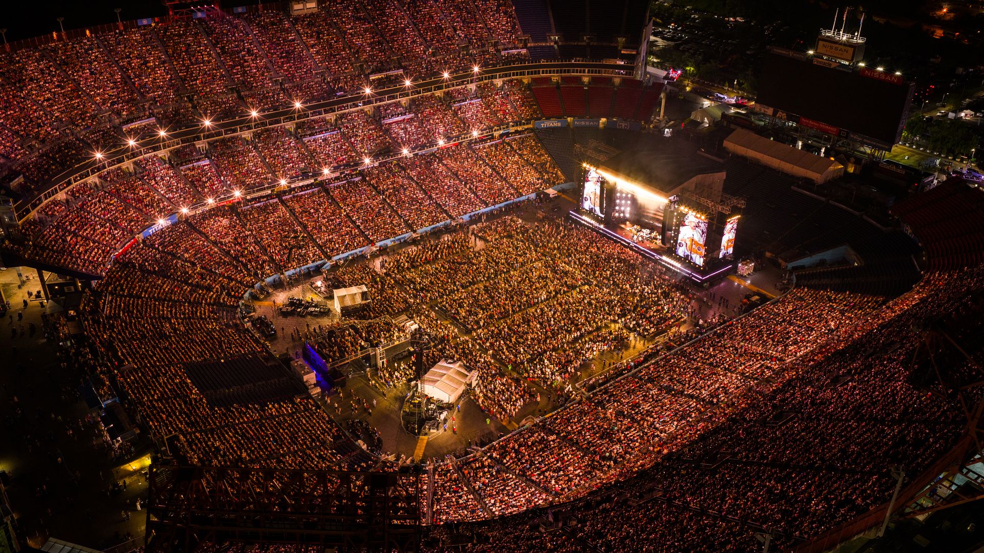 Overhead night view of a packed outdoor stadium concert. A bright stage with large screens and flames on the right, crowd filling all stands, illuminated by stadium lights.