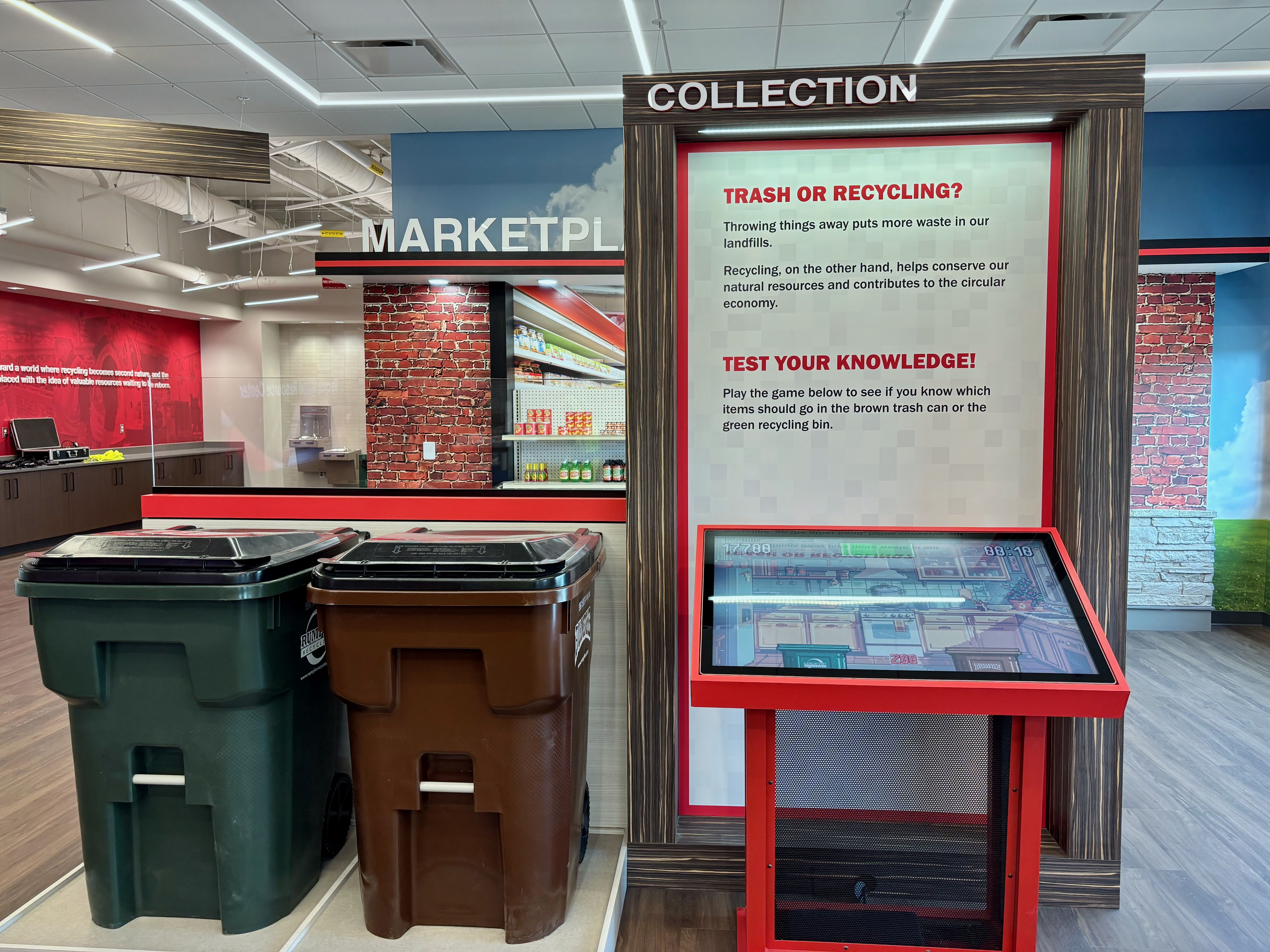 Inside a store display, a tall wood-framed panel reads "COLLECTION" beside a poster titled "TRASH OR RECYCLING?" about waste, with green and brown trash bins upfront, brick walls, and a red kiosk.