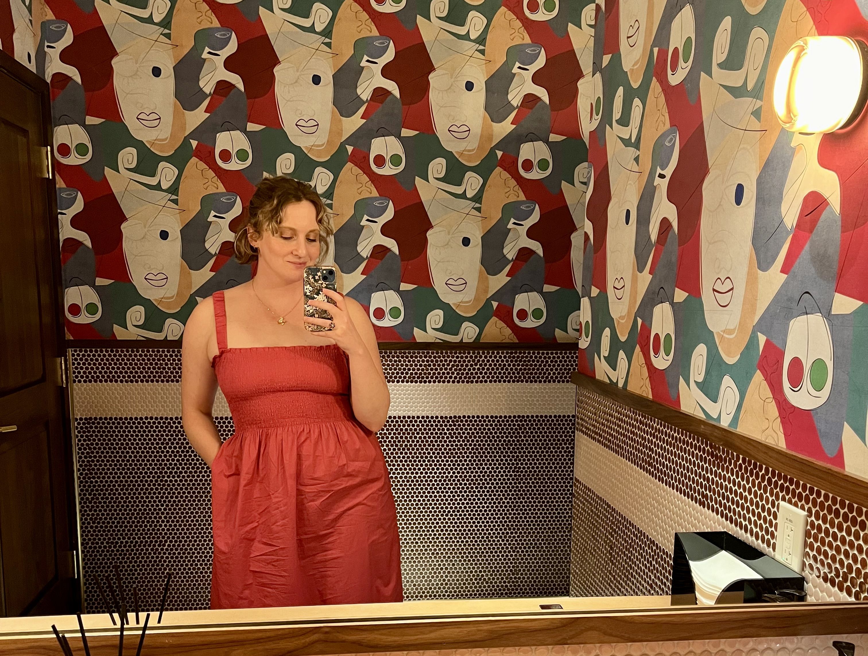 A woman in a pink dress taking a selfie in the mirror of a bathroom with multi-colored abstract face wallpaper.