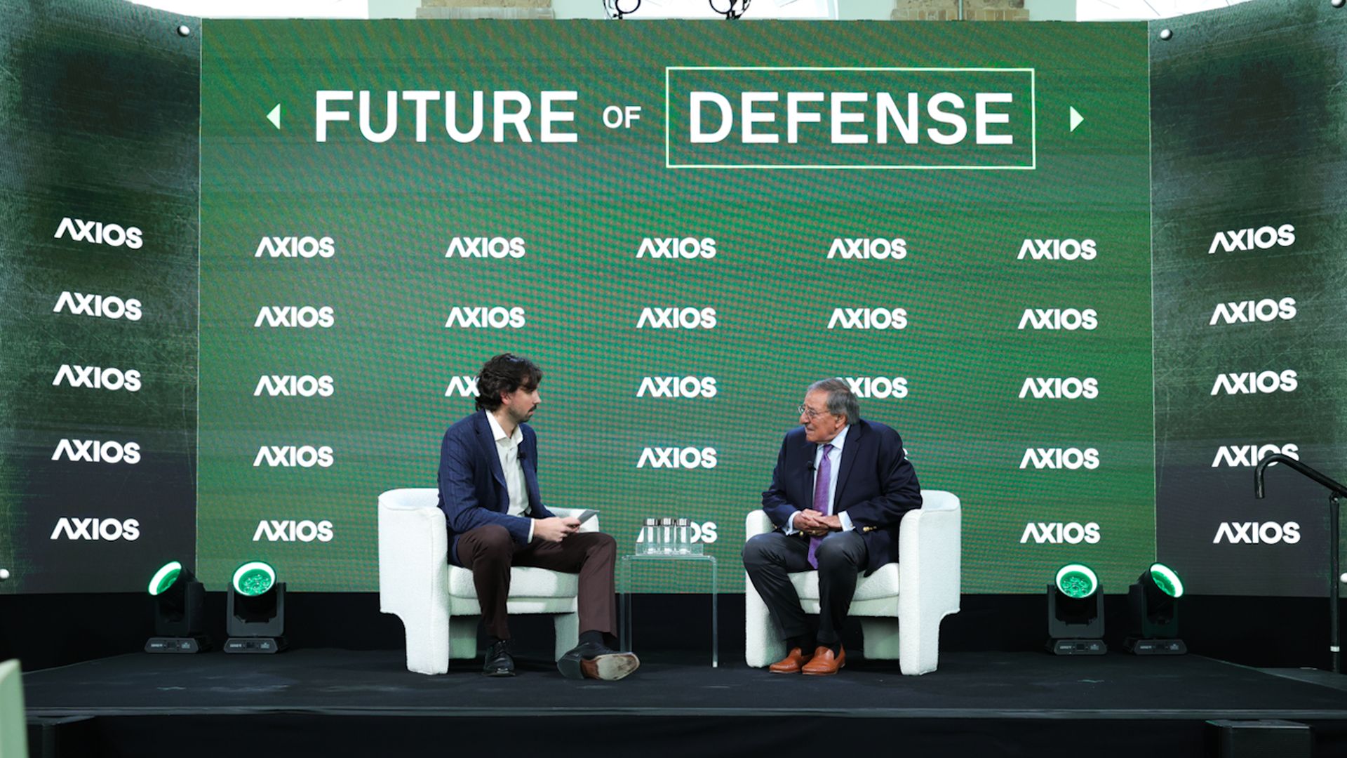 Two men seated on white chairs on a stage with a green backdrop displaying "FUTURE OF DEFENSE" and multiple "AXIOS" logos, engaged in conversation during an event.