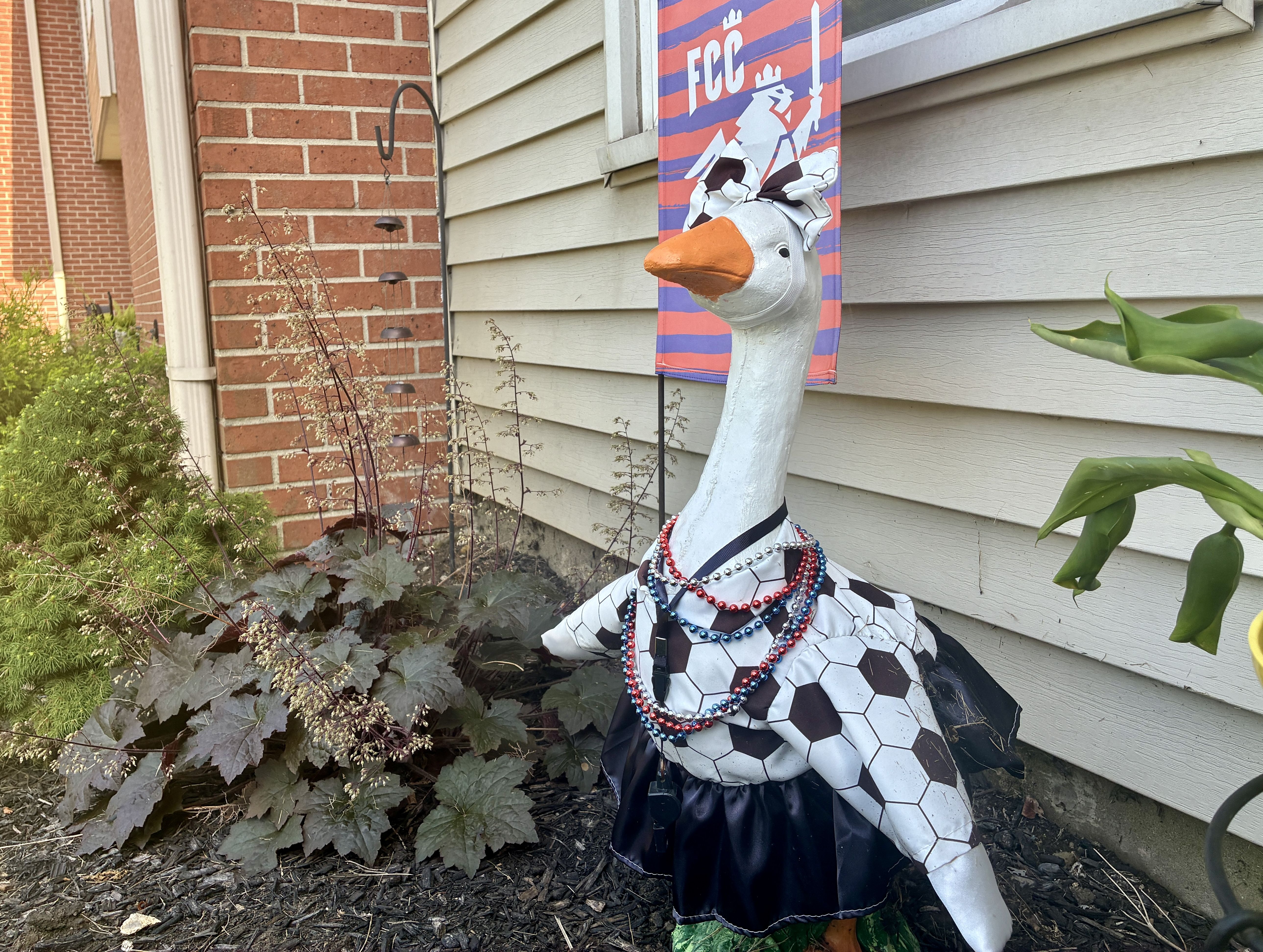 Decorative white goose statue in a black-and-white dress with orange beak, red and blue bead necklaces, and a headscarf, placed outside beside a beige clapboard house with a banner behind.