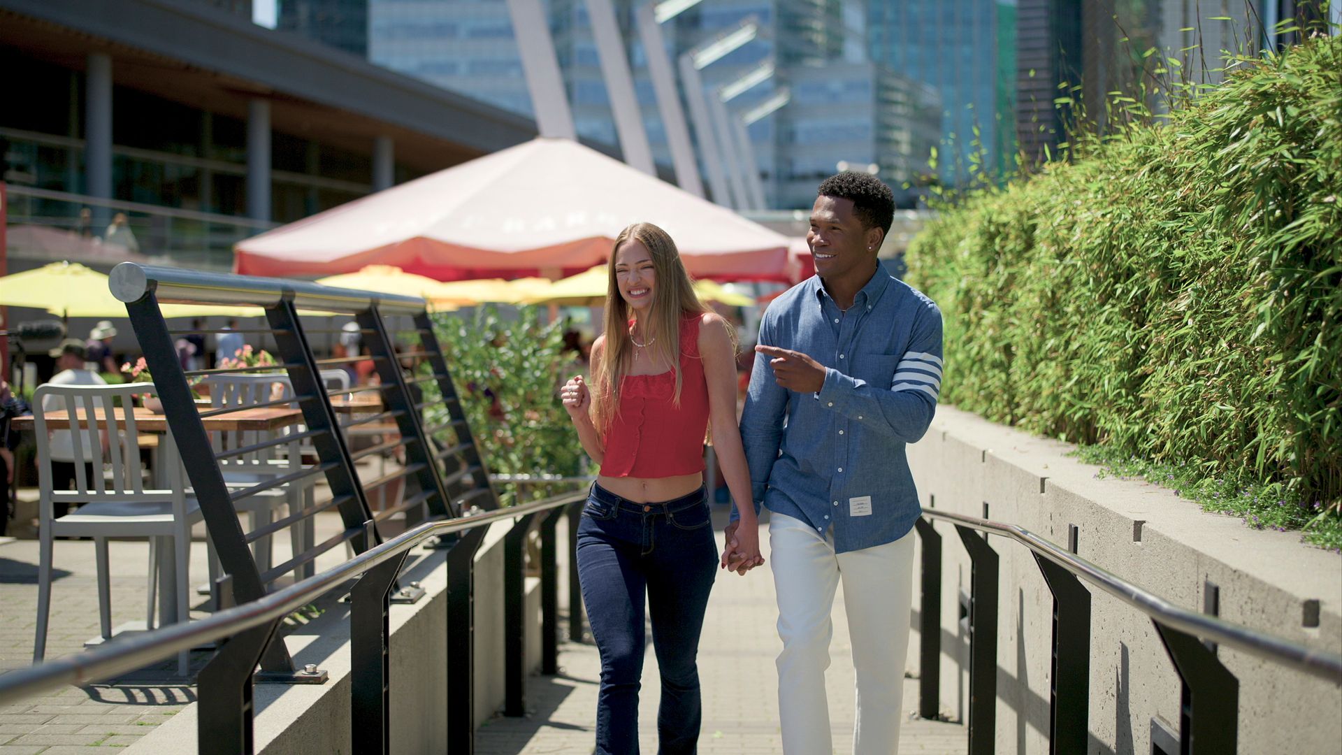 A couple walking hand-in-hand along a sunny urban promenade. She wears a red sleeveless top; he wears a blue shirt and white pants. 
