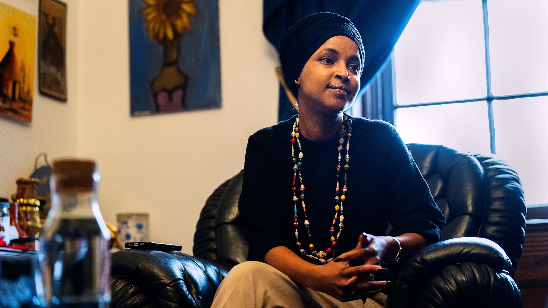 A woman wears a black head covering, a colorful beaded necklace and a black shirt (Ilhan Omar) as she sits in a black chair in front of a window and artwork of a flower.
