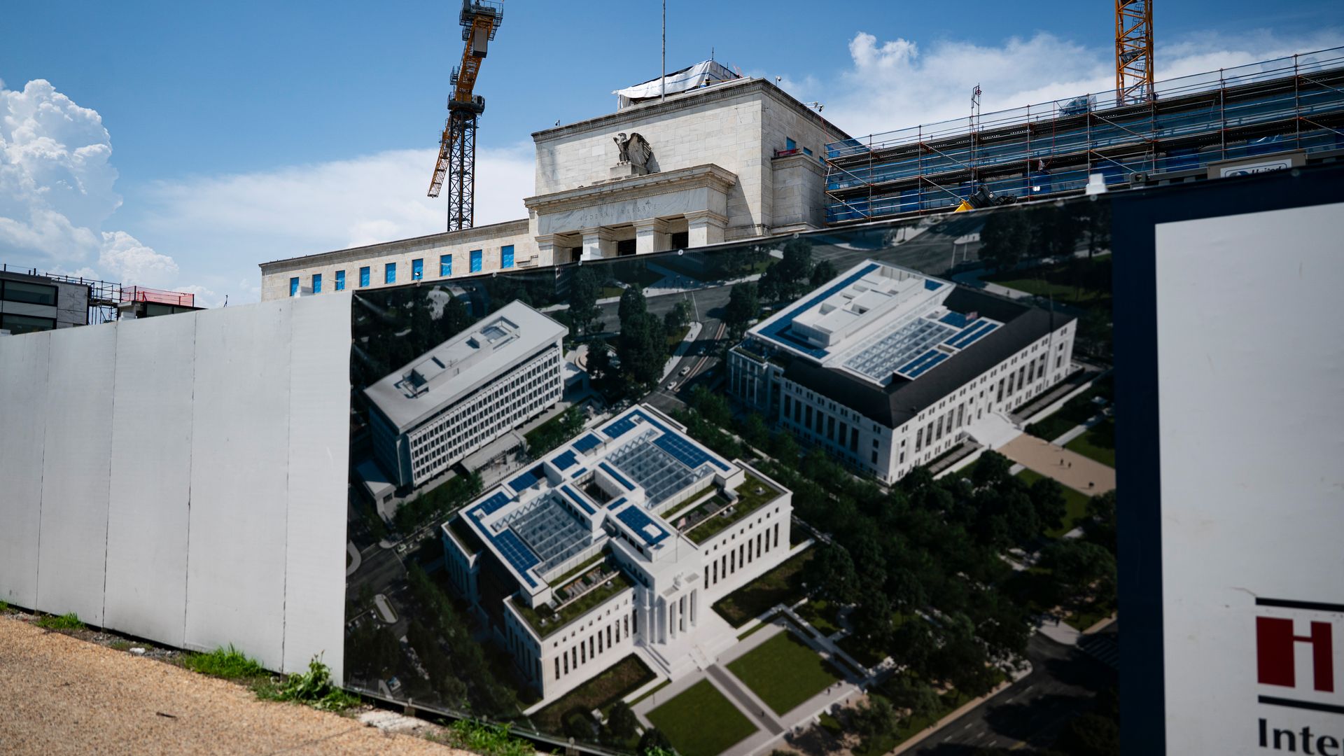 Construction site with white fence displaying a large image of planned buildings. Behind the fence is an older stone building under renovation with cranes against a blue sky.