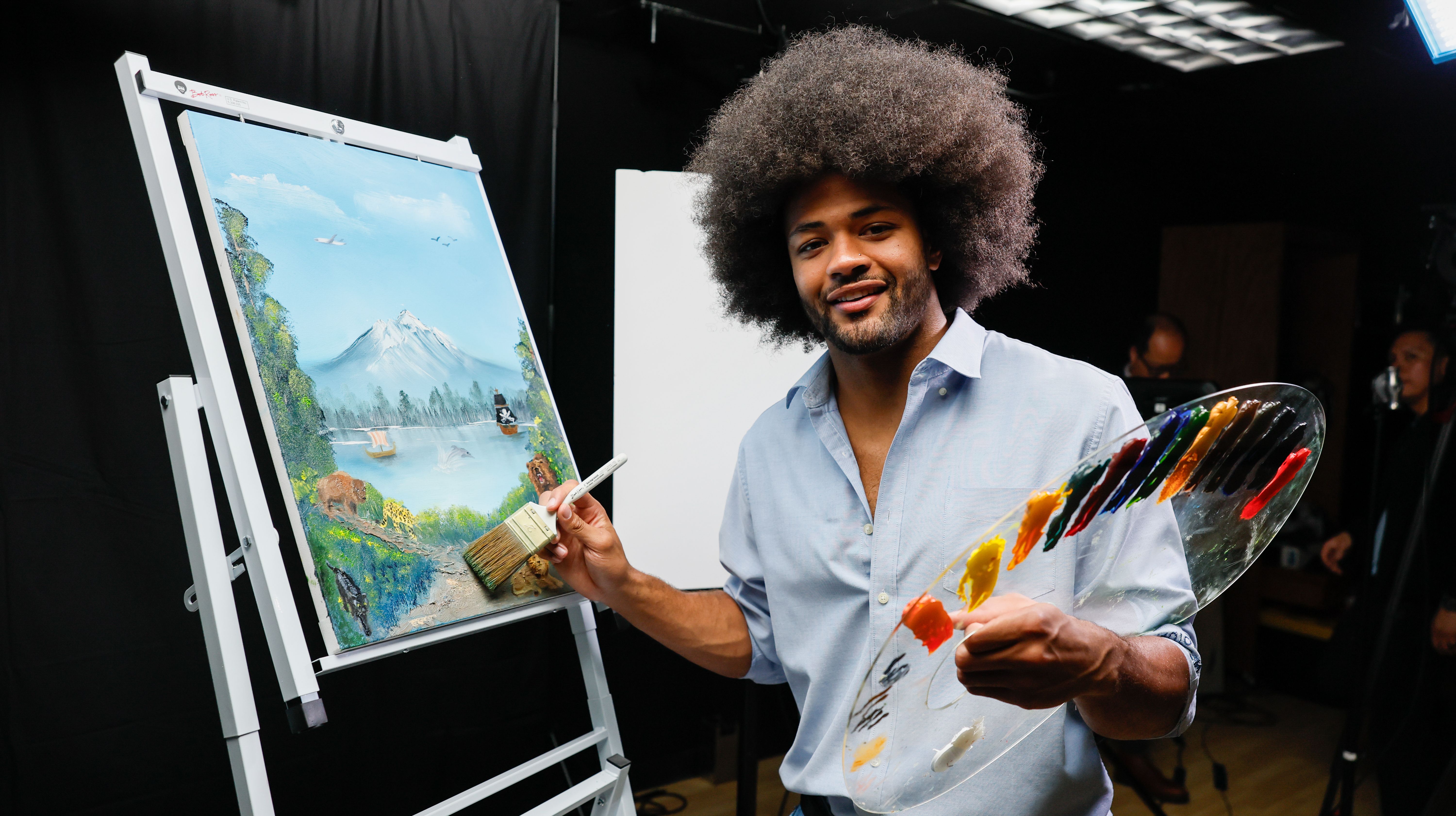 A man with a large afro and a light blue shirt paints a landscape on an easel, featuring a mountain, lake and trees; he holds a brush and a painter's palette.