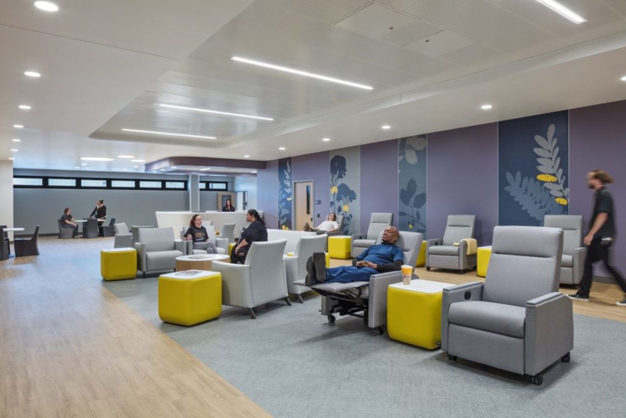 Modern medical center lounge with purple accent walls and floral blue panels, gray recliners, and bright yellow ottomans. People relax and chat; a man lounges in a chair as a receptionist desk appears in the background.