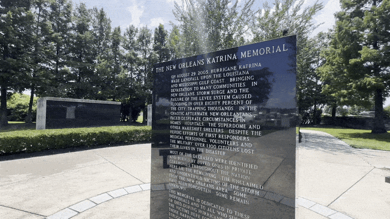 Black stone monument at the New Orleans Katrina Memorial with engraved text about Hurricane Katrina's impact, surrounded by trees and greenery on a paved area under a blue sky.