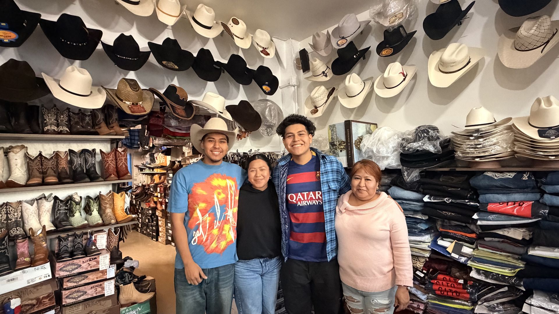 Four people pose together in a hat shop, smiling at the camera. The store is crowded with cowboy hats on the ceiling and walls, plus boots, shirts, and folded goods stacked around them.