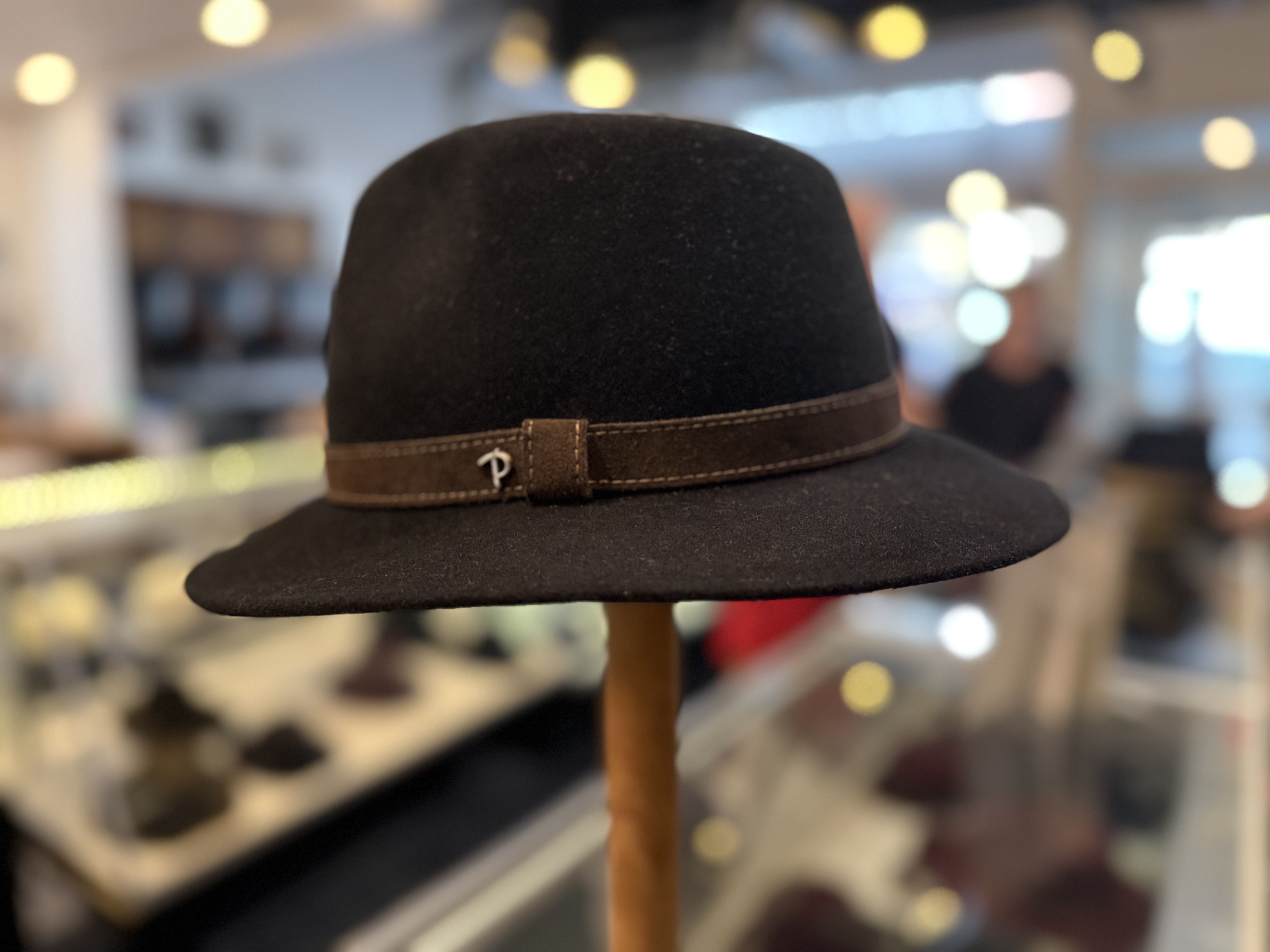Close-up of a black felt hat with a brown leather band and a small metal pin on a hat stand, against a blurred store interior with warm yellow bokeh lights in the background.