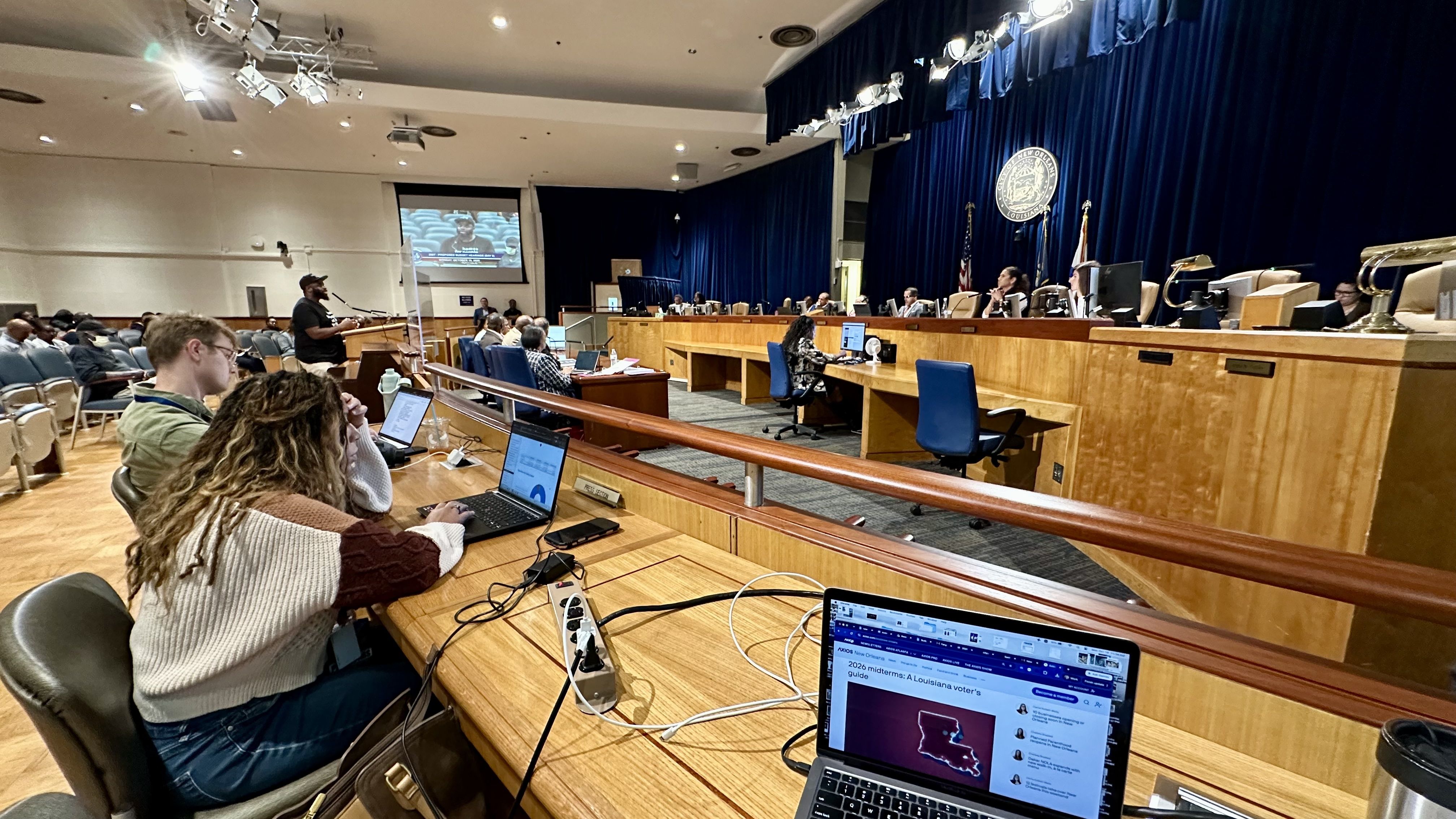 Wide-angle view of a government hearing: officials sit at a long wooden dais, blue curtains with a state seal, flags, a speaker at the podium, and attendees using laptops.