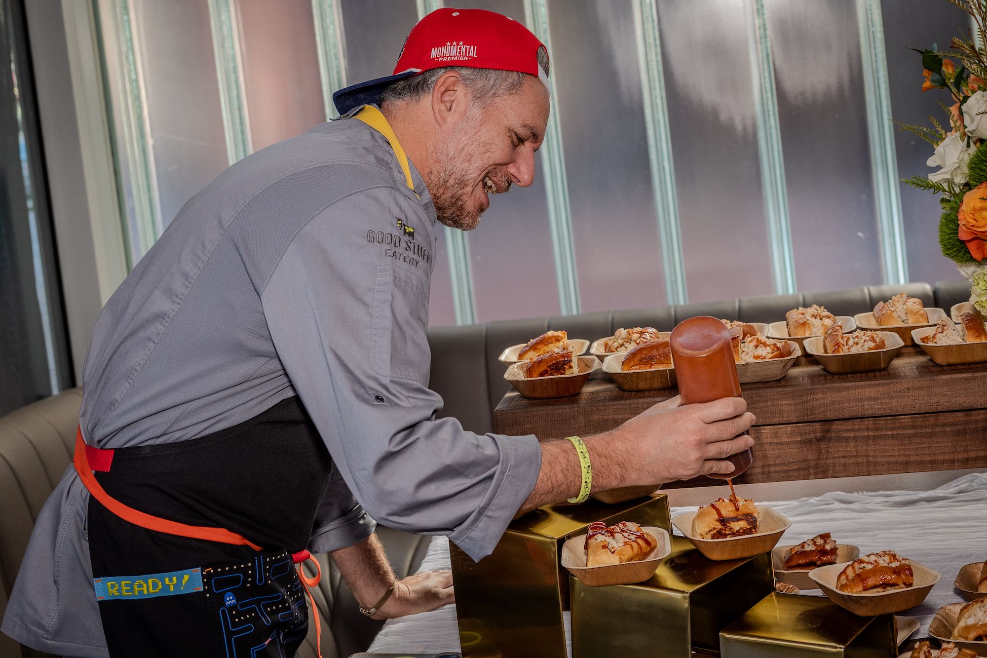 Spike Mendelsohn wearing a red cap, gray chef coat, and black apron is smiling while drizzling sauce over food in trays on a wooden table with a white cloth and gold stands.