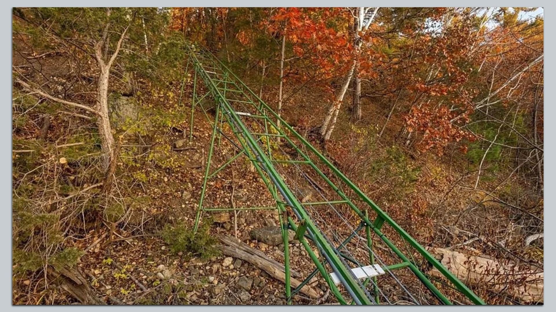 Green metal roller coaster track running diagonally through a forest with autumn foliage in orange, red, and green hues.