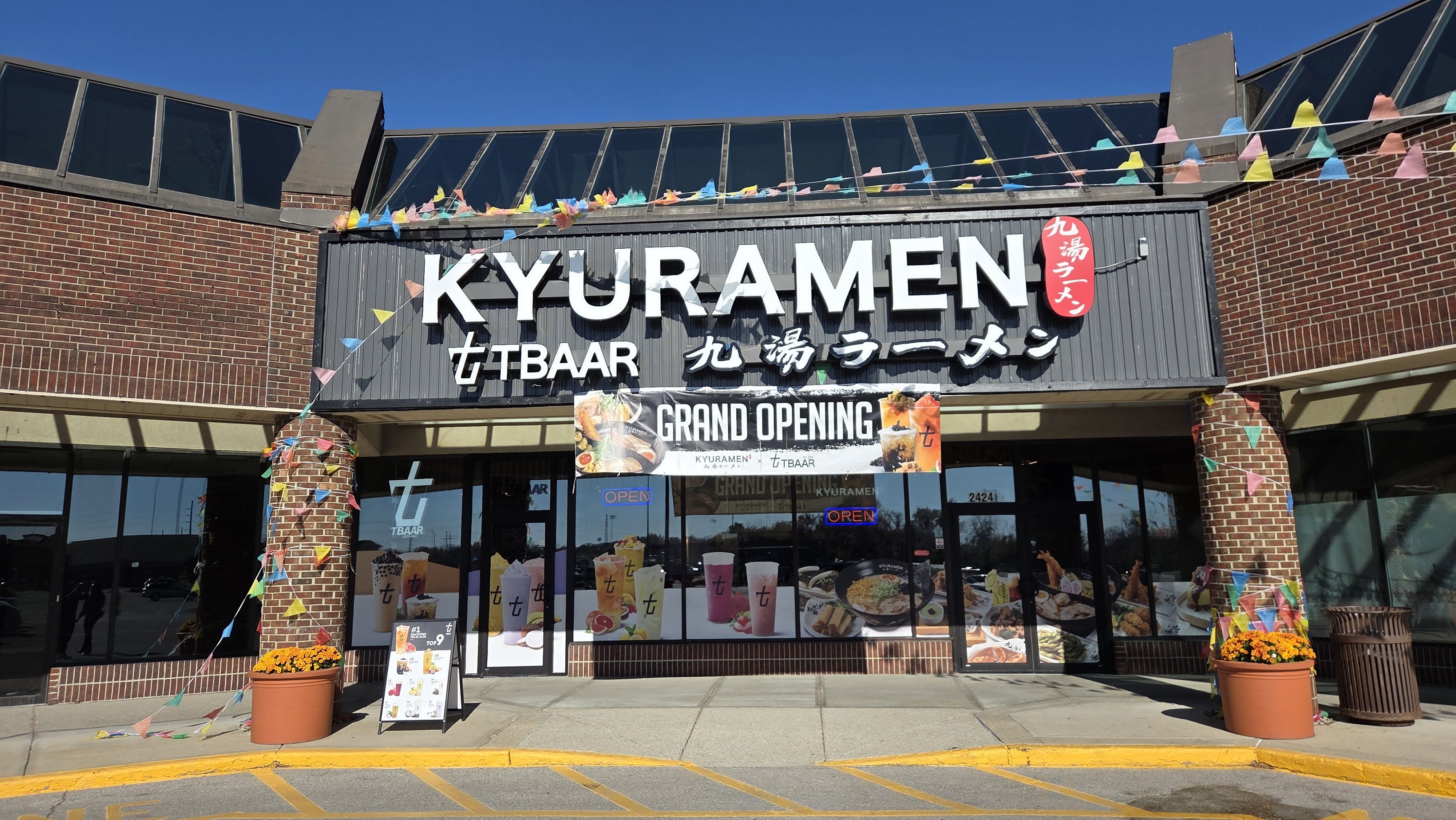 Exterior of KYURAMEN tTBAAR restaurant with brick walls, colorful triangular flags, orange flowers in pots, a "GRAND OPENING" banner, and bright blue sky.