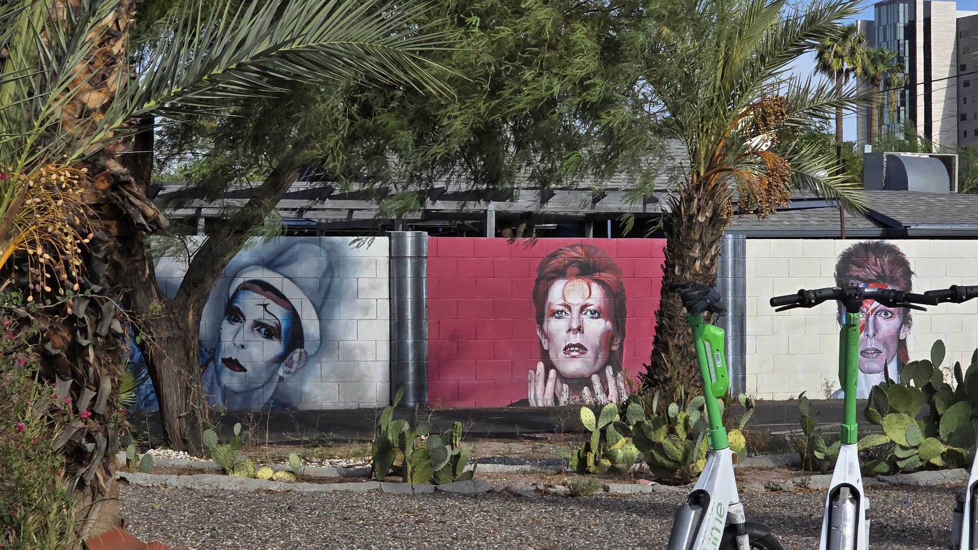Sunlit outdoor scene with a muraled cinder-wall showing large portraits in blue and red tones, palm trees, desert plants, and a row of green-and-white electric scooters in the foreground.