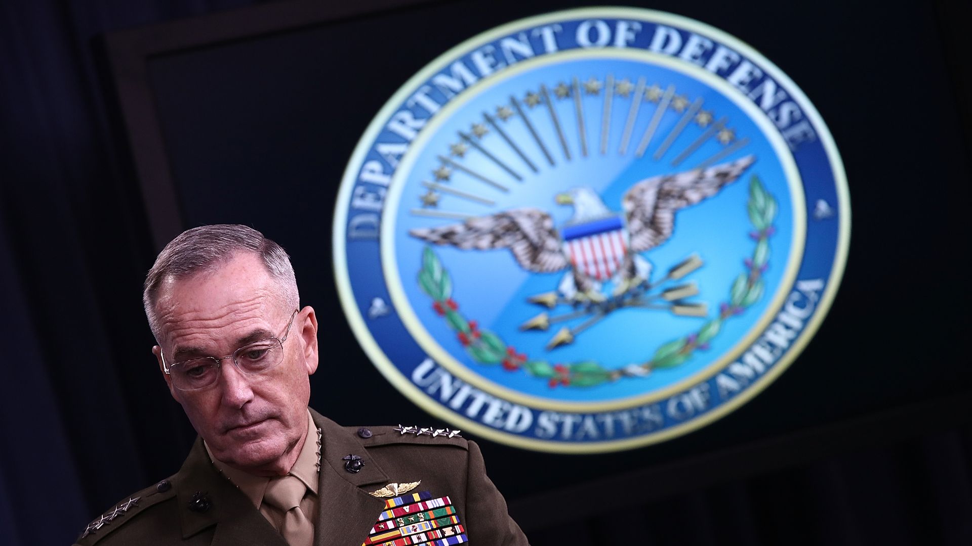 A man in a decorated military uniform stands in a dark room. The Defense Department logo is seen over his shoulders, bright and blue.