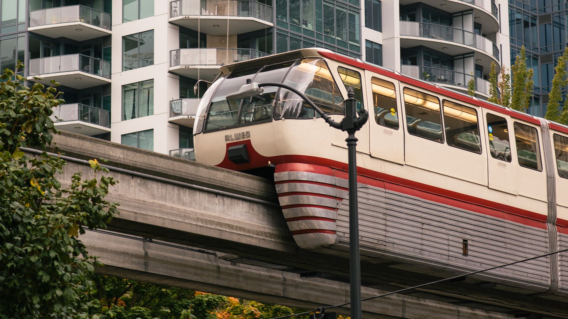 A vintage red and white monorail train on an elevated track in a city with modern high-rise glass buildings and green trees below on a cloudy day.