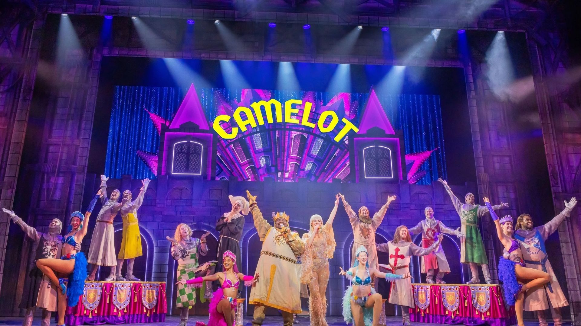 Colorful theater stage featuring a large "Camelot" sign; a diverse cast in medieval and showgirl costumes pose with arms raised before a castle backdrop, bathed in blue and purple lights.