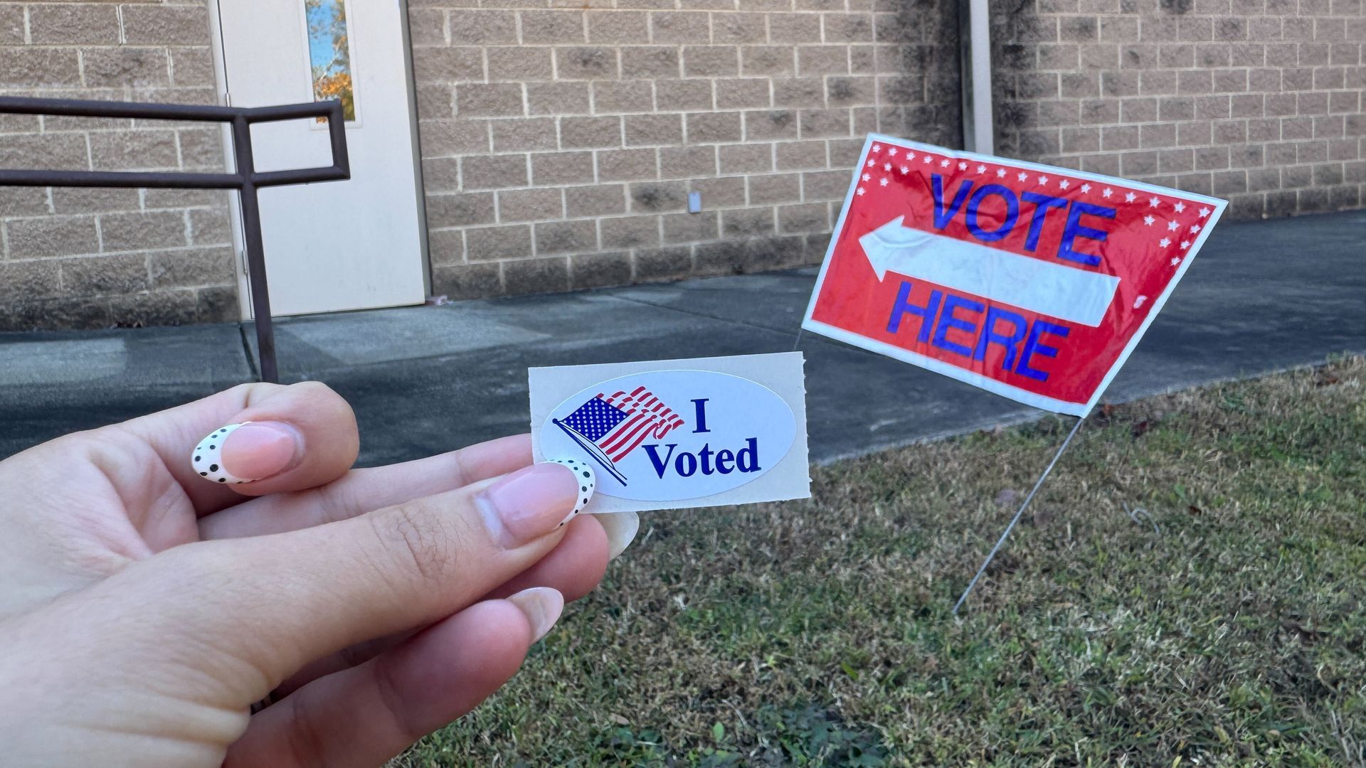 Person holding an "I Voted" sticker near a brick building with a door and red "Vote Here" sign pointing left on grass.